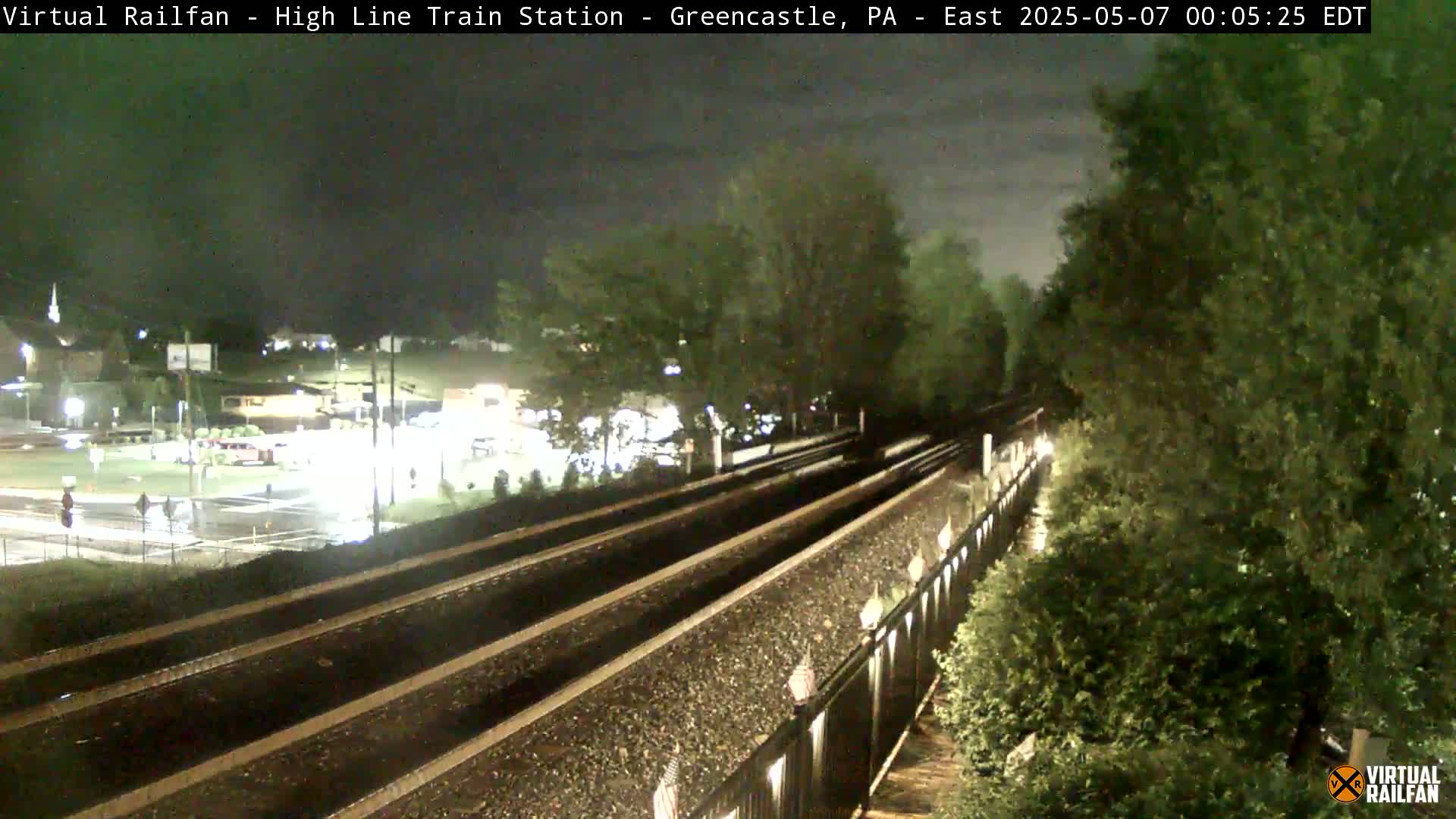 At night, a view of train tracks extends from a slightly elevated vantage point, passing through a well-lit area with buildings and greenery visible in the background.