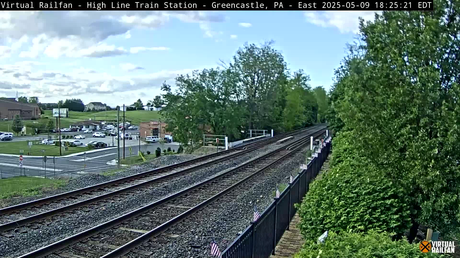 A partly cloudy day shows a view of railroad tracks extending into the distance, passing by a parking lot and some buildings, alongside lush green vegetation.