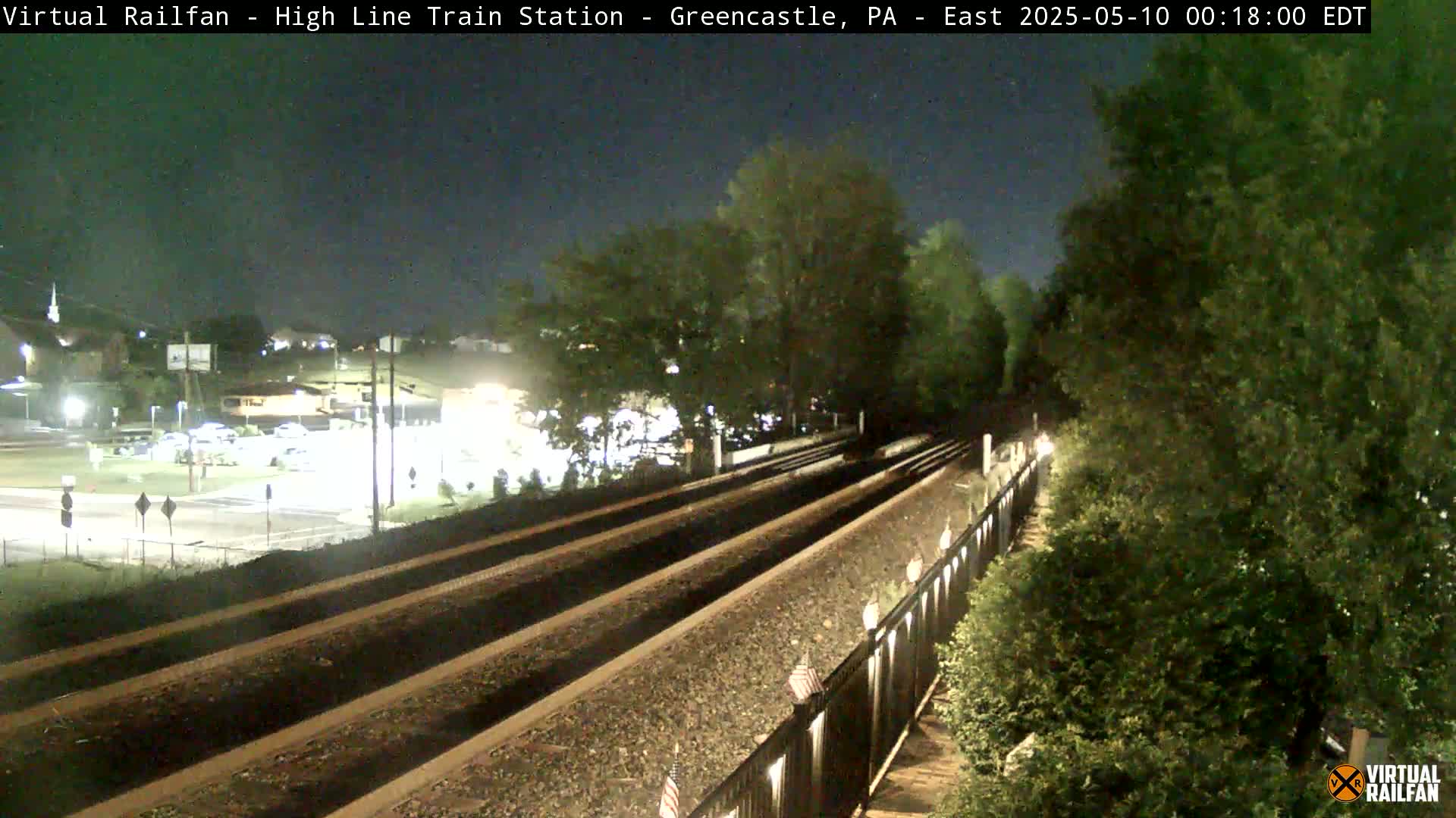 At night, two sets of train tracks stretch into the distance, bordered by lush green vegetation and a black fence, with a road and buildings faintly visible in the background.