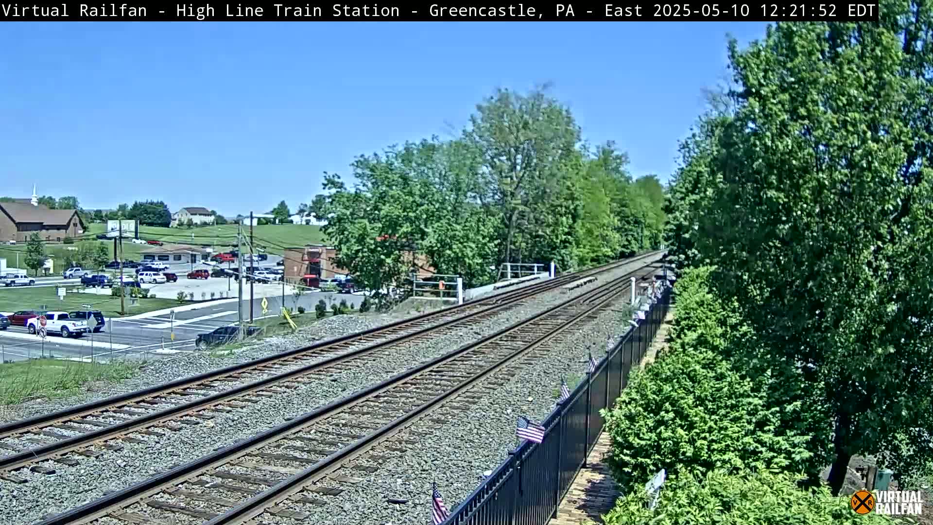 Under a clear blue sky, a long stretch of train tracks extends into the distance, passing through a lush green landscape with buildings and parked cars visible in the background.