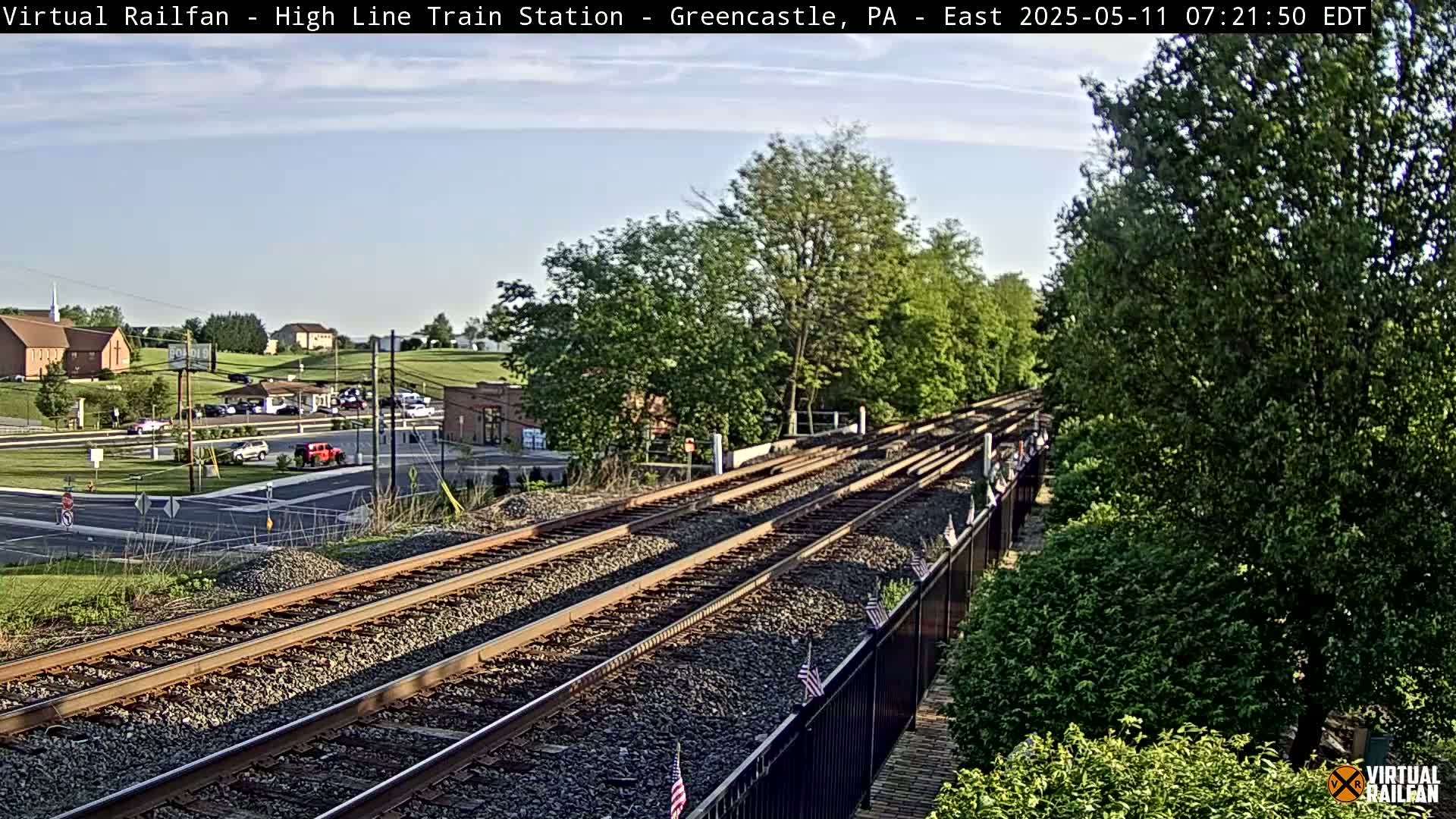 Train tracks extend into the distance under a partly sunny sky, passing alongside a road with cars and buildings, and bordered by lush green trees and a black fence.