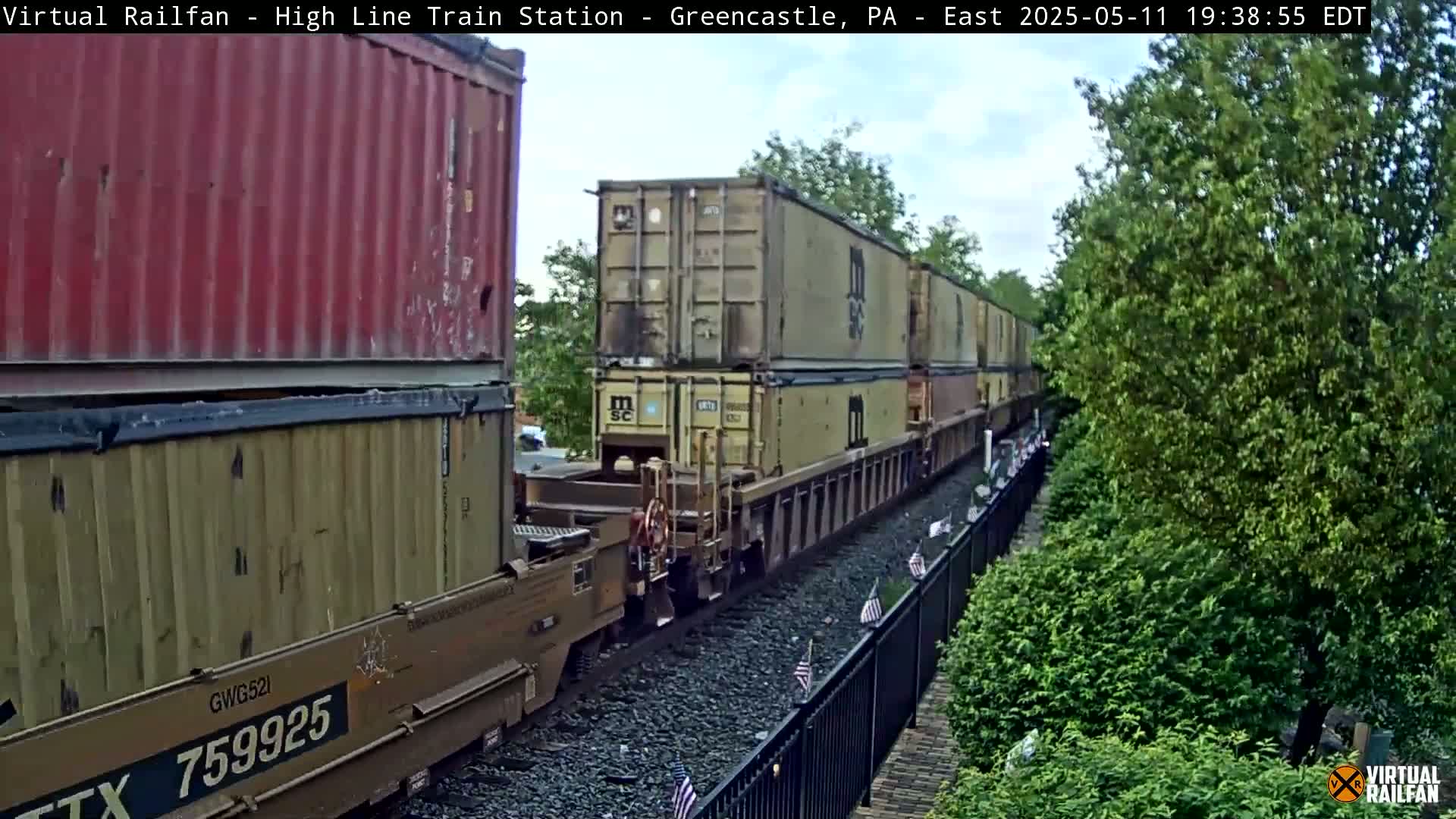 A freight train carrying stacked shipping containers moves eastward along a track beside a lush green landscape under a partly cloudy sky.