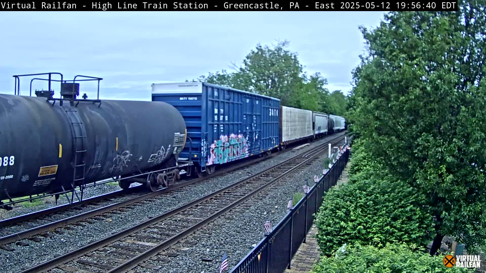 A freight train, consisting of several tank cars and boxcars, some with graffiti, travels along a railway track beside lush green vegetation under a partly cloudy sky.