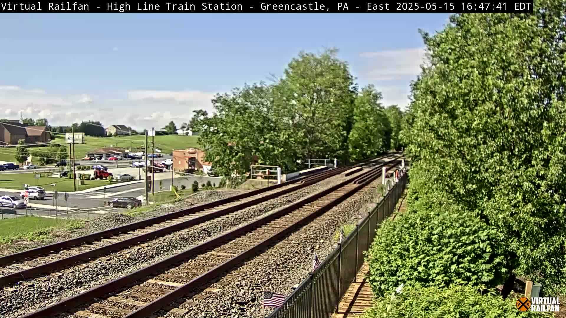 A view of railroad tracks extending into the distance, alongside lush green vegetation under a partly cloudy sky.