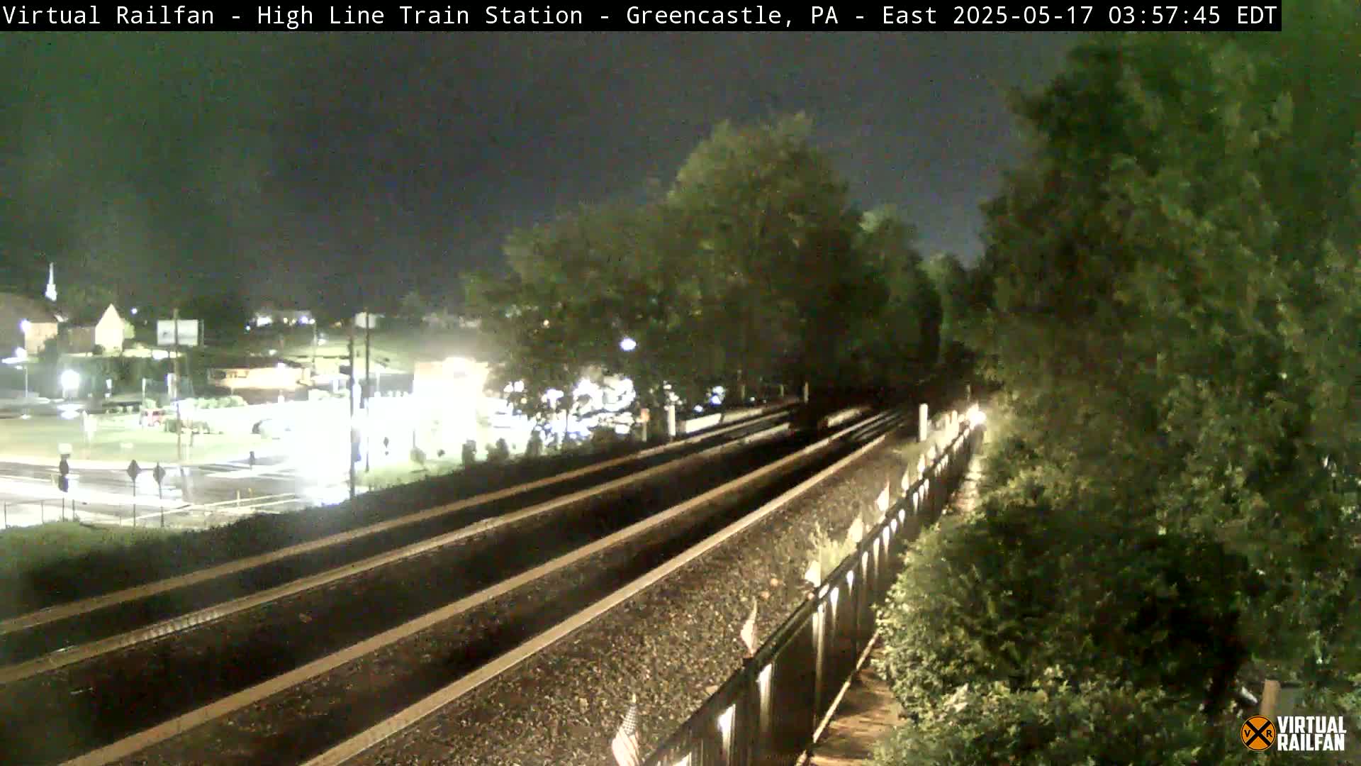 At night, three sets of train tracks extend into the distance, flanked by lush green vegetation and a metal fence, with a dimly lit intersection visible in the background.