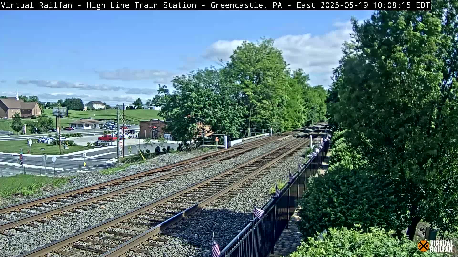 A view of railroad tracks extending into the distance, with lush green trees and buildings visible in the background under a partly cloudy sky.