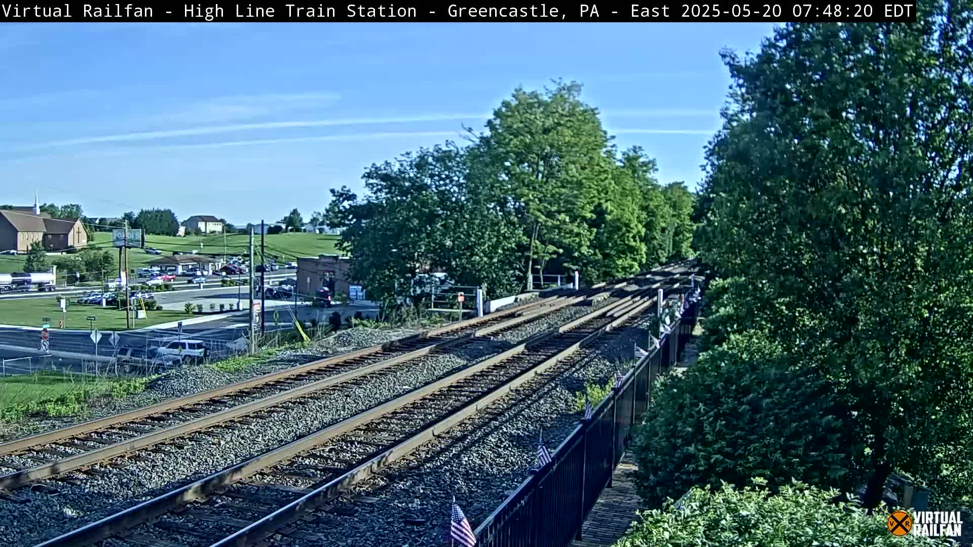 A sunny day view of a railway track, with multiple train lines extending into the distance, passing alongside a road, a parking lot, and some buildings.