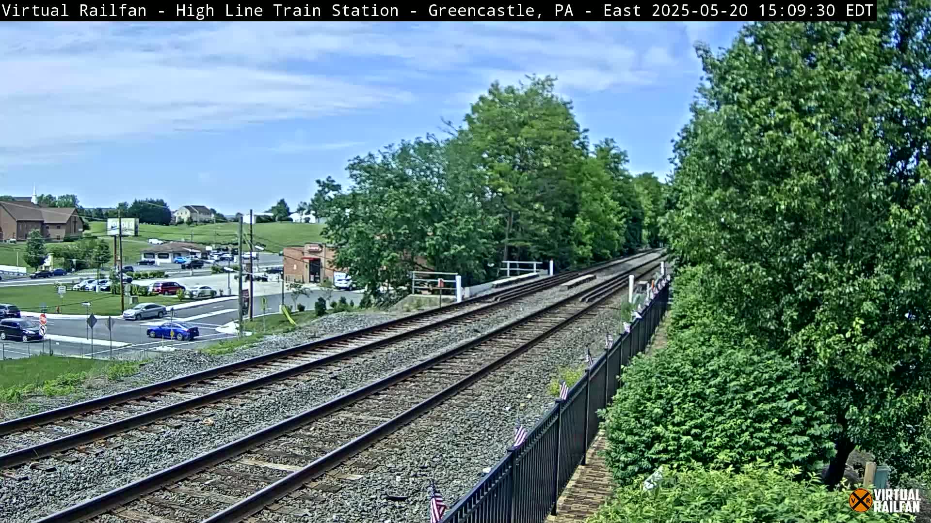 A view of train tracks extending into the distance on a partly sunny day, with cars parked in a lot and buildings visible in the background.