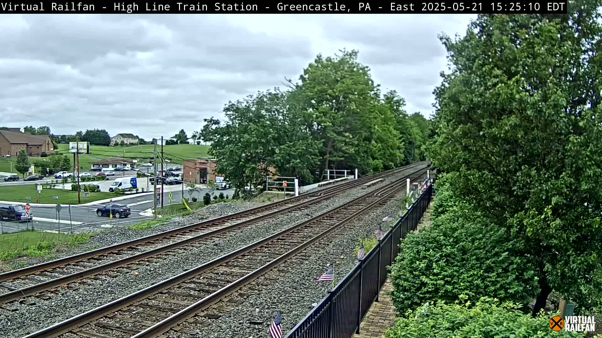 A view of train tracks extending into the distance under a partly cloudy sky, alongside a road with cars and buildings visible in the background.