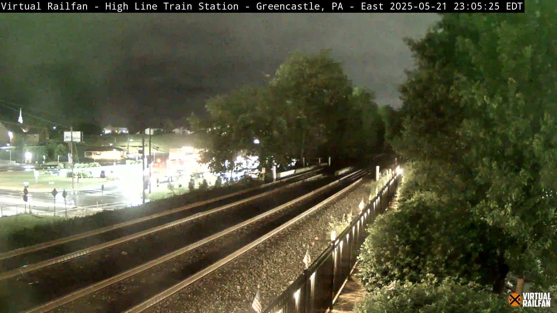 A nighttime view of a rain-slicked train track splitting into two paths, surrounded by lush green vegetation under a dark, cloudy sky.