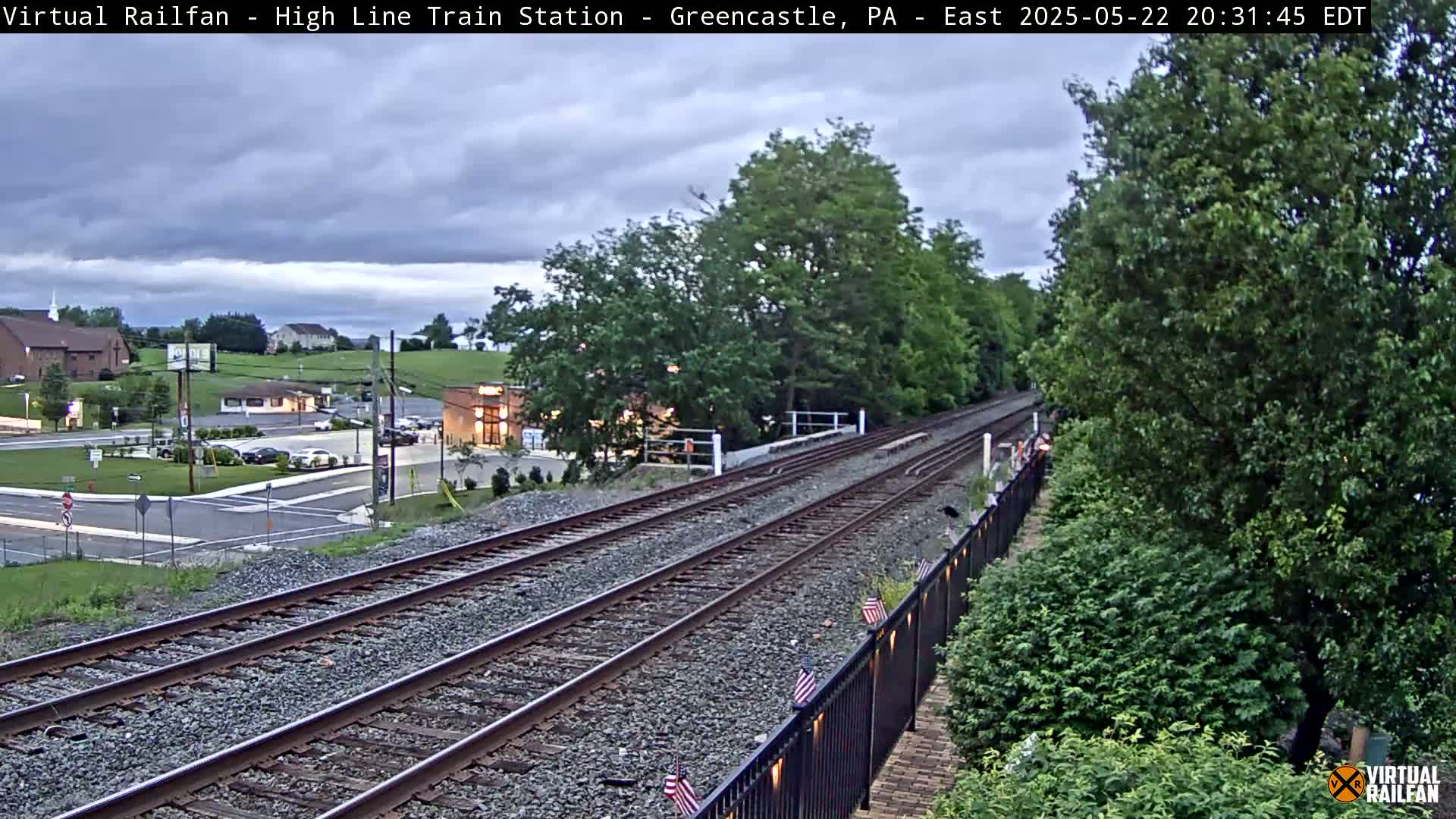 A view of railroad tracks stretching into the distance under an overcast sky, alongside a roadway with buildings and greenery.