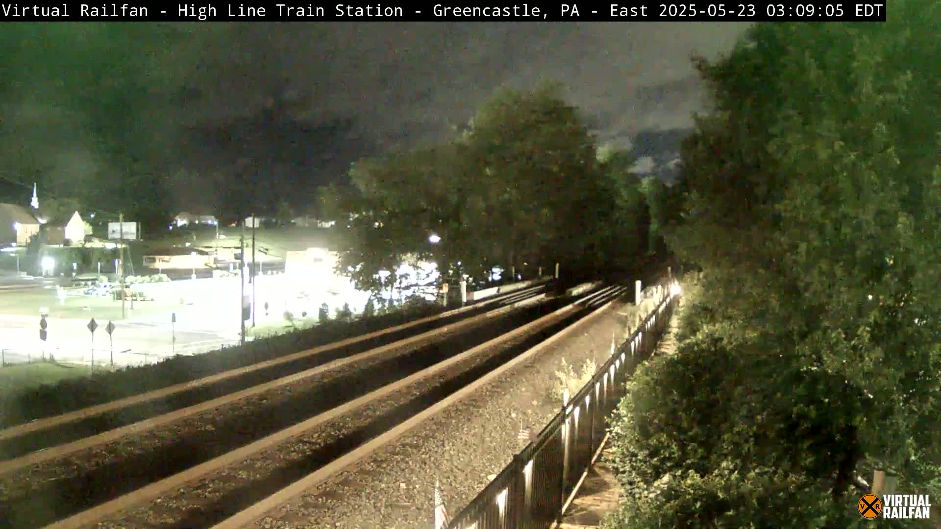 A nighttime view of train tracks extending into the distance, bordered by lush green vegetation, under a dark sky.