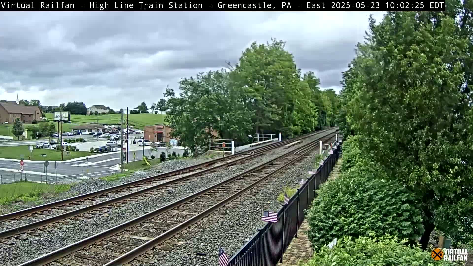 A view of train tracks extending into the distance, under a partly cloudy sky, with lush green trees and foliage lining the tracks and a parking lot visible in the background.