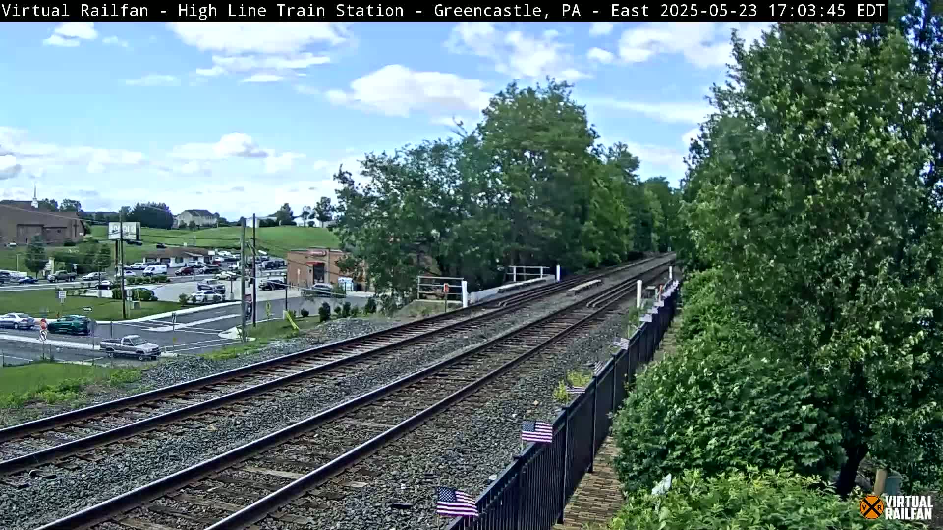 A high-angle view of a train track on a partly cloudy day, with a parking lot and buildings visible in the background.