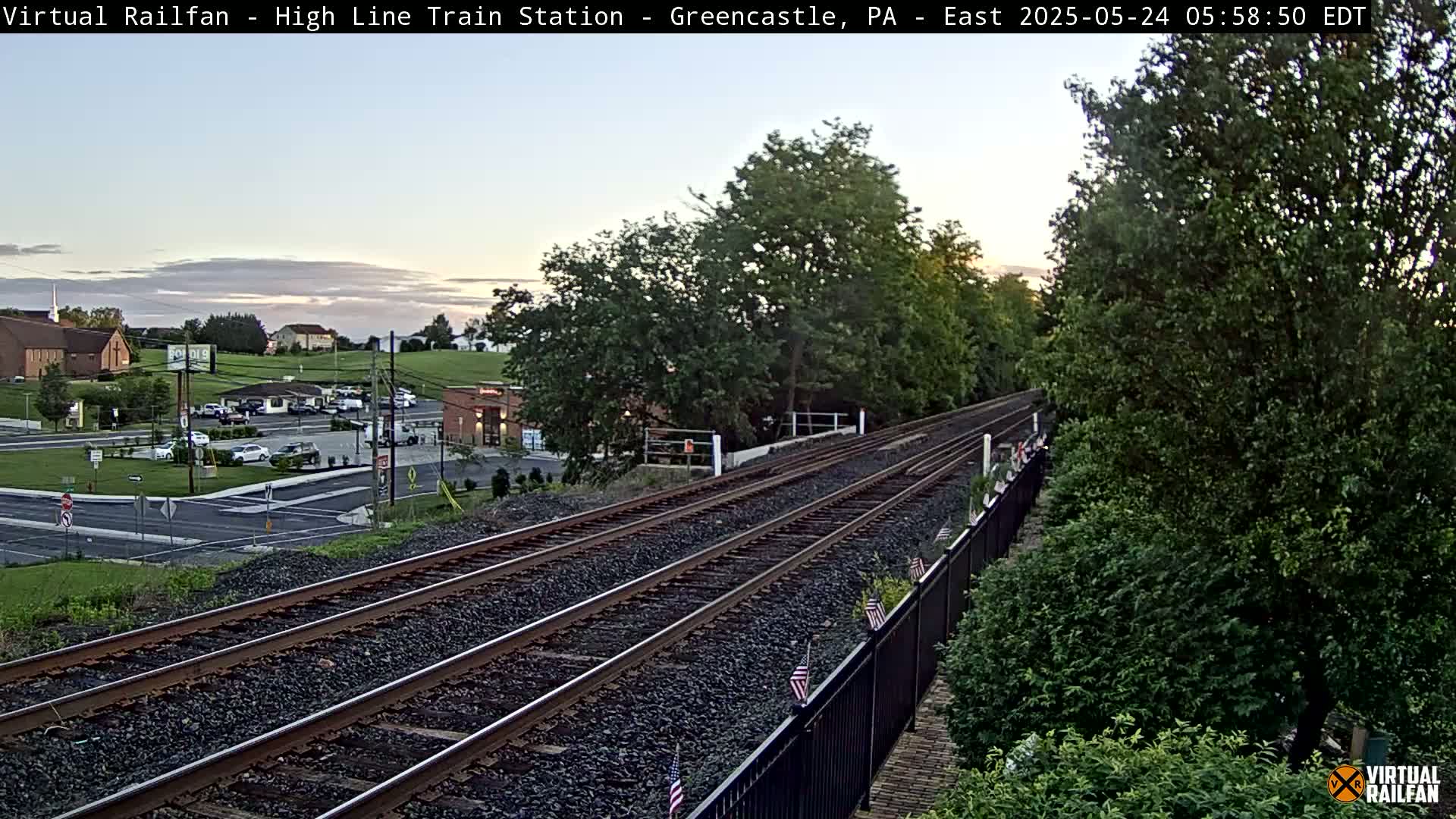 A twilight view of a railway track extending into the distance, bordered by lush green trees and a small town visible in the background.