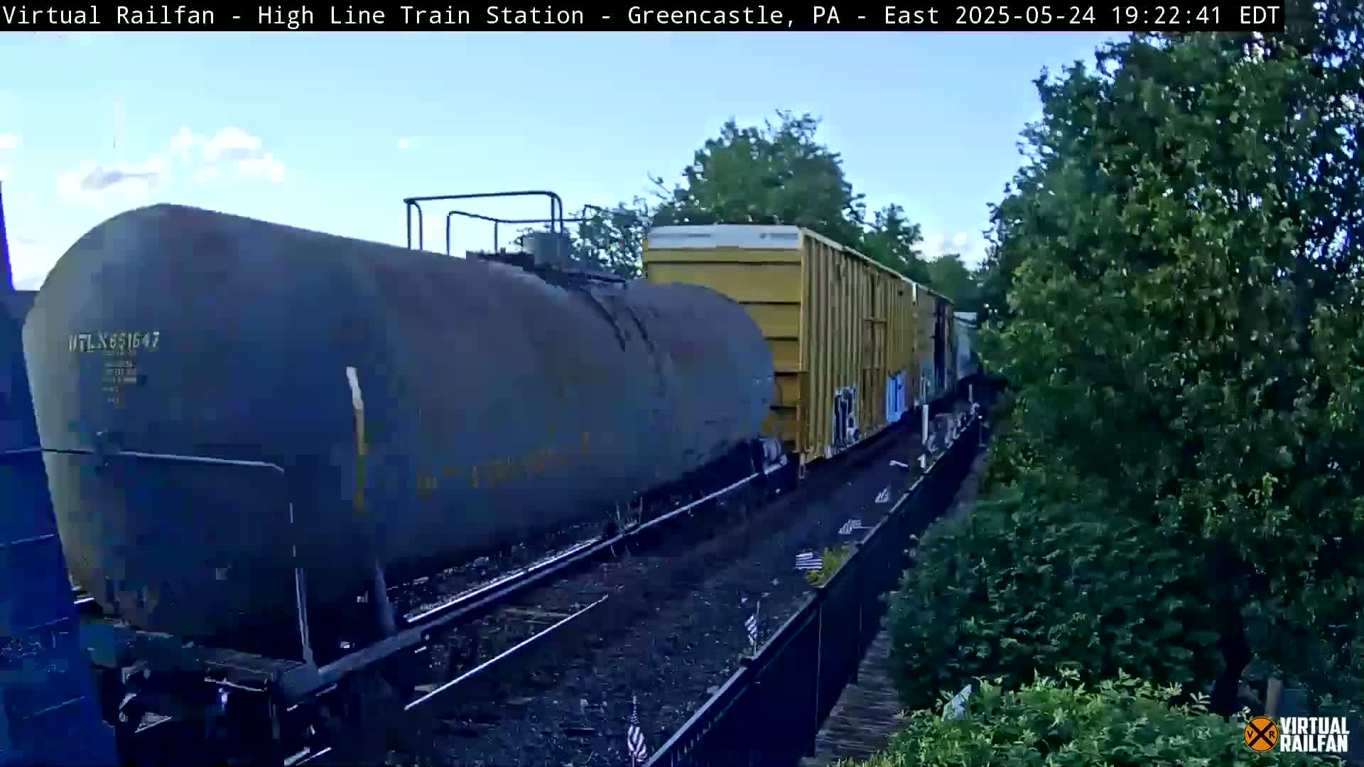 A freight train, including a large tanker car and boxcars, passes by under a partly cloudy sky.