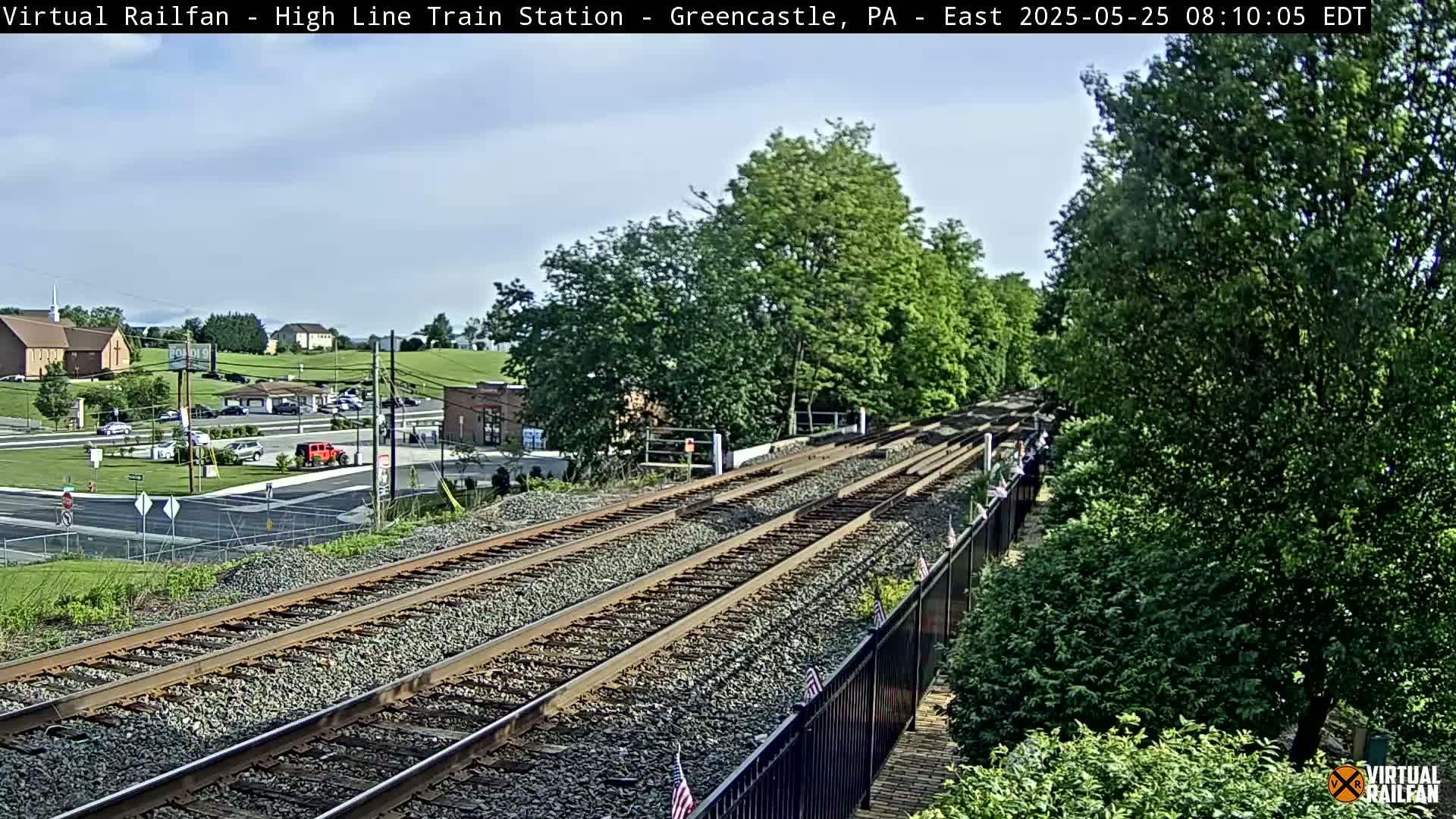 A mostly sunny view of a train track elevated above a roadway and small town, with lush green trees and shrubs visible alongside the tracks.