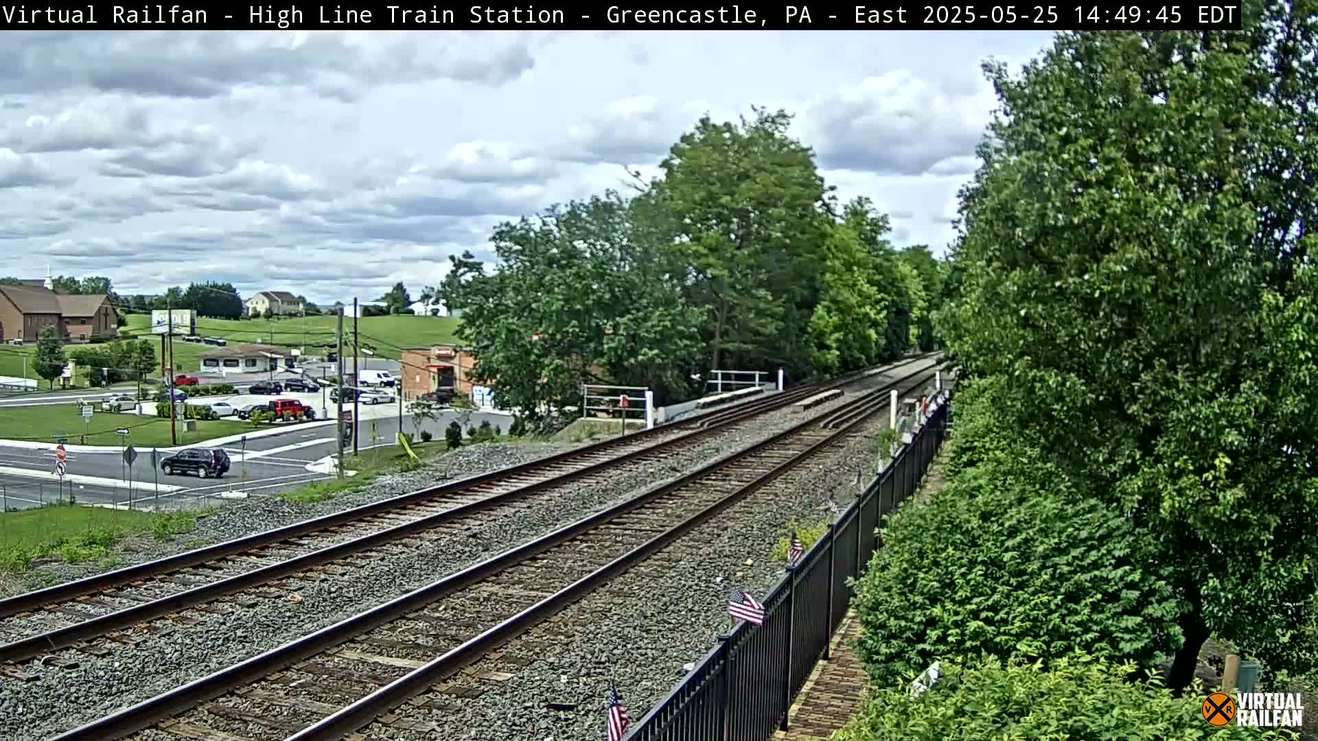 A partly cloudy day shows train tracks extending from a small town into a lush green landscape.