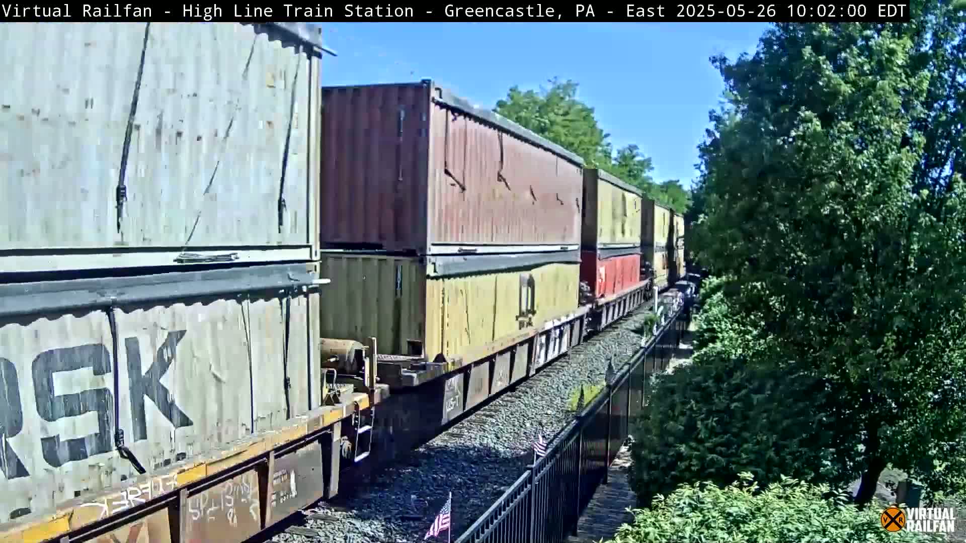 A long freight train carrying multiple shipping containers moves eastward on a sunny day.