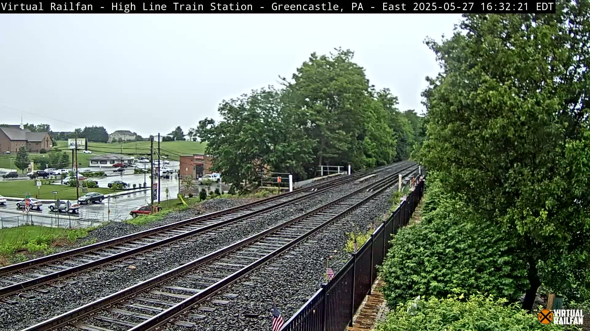 A view of railroad tracks extending into the distance on a slightly overcast day, with green trees and a town visible in the background.