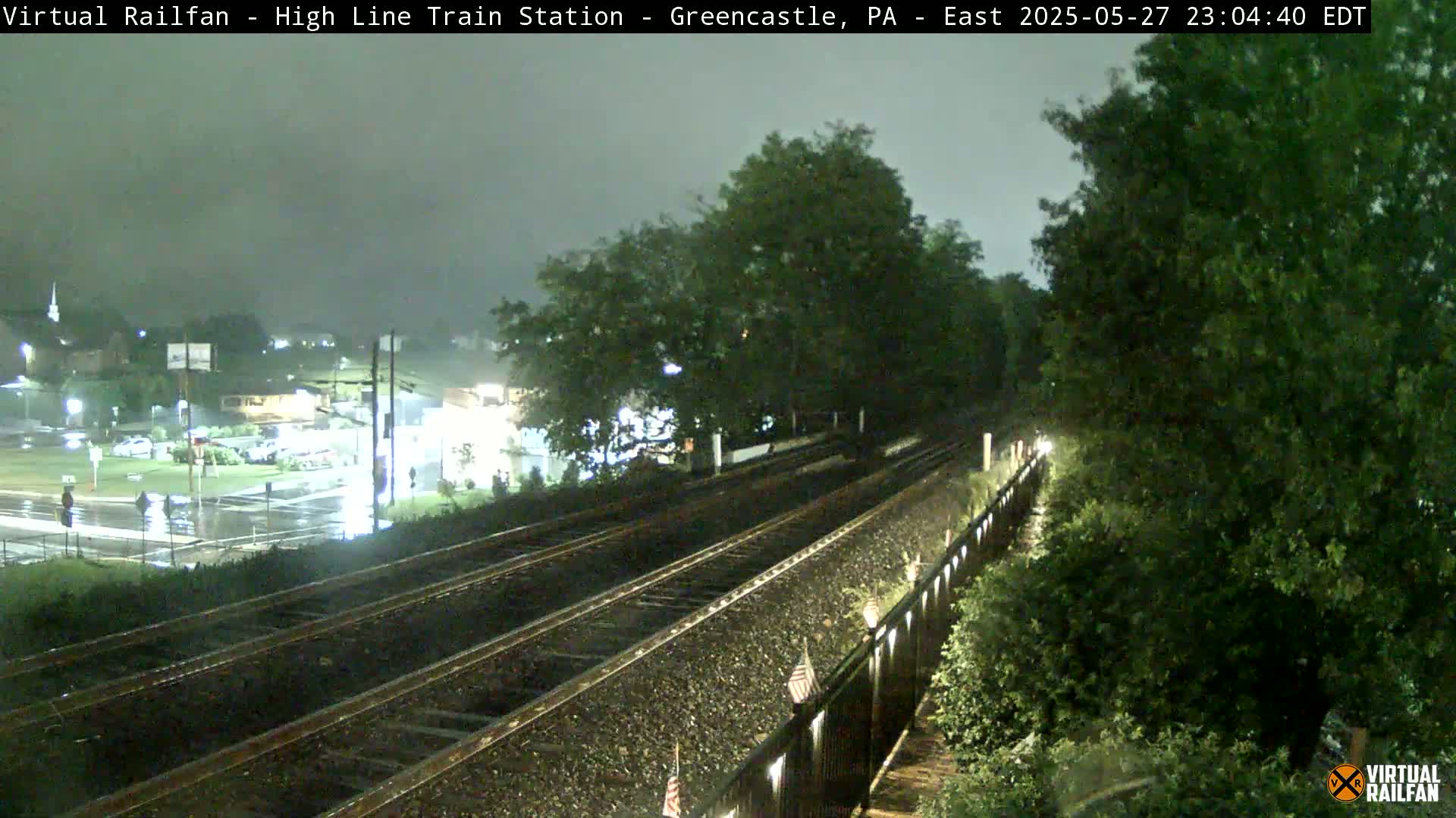 A nighttime view of rain-slicked train tracks extending into the distance, flanked by lush green vegetation under a dark, overcast sky.