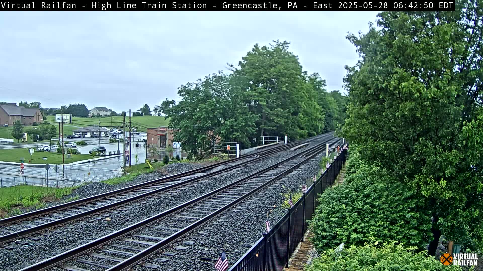 A view of double train tracks extending into the distance, bordered by a black fence, lush green vegetation, and a parking lot with cars visible in the background under an overcast sky.