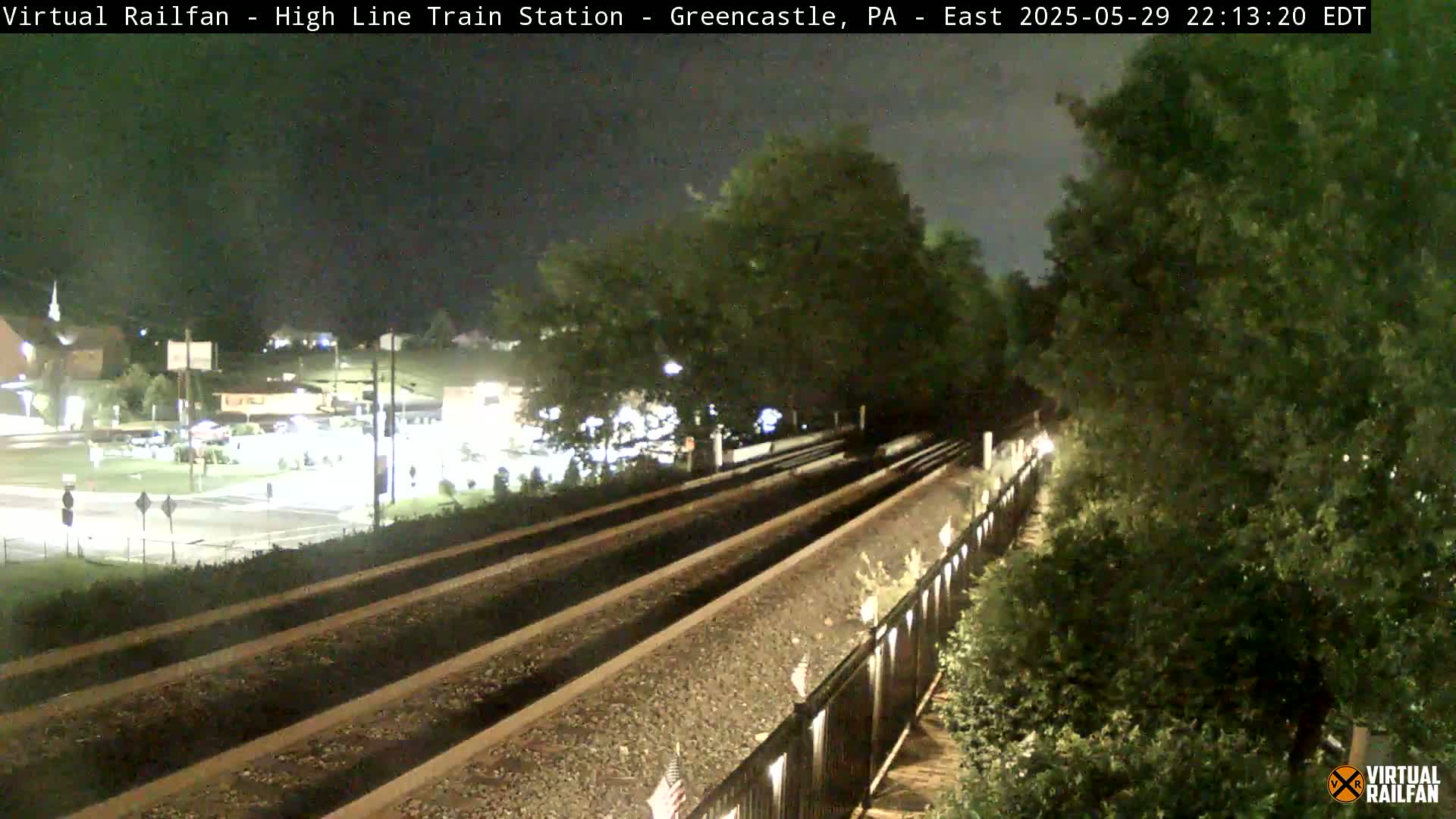 A nighttime view of a train track, bordered by a fence and lush greenery, with a dimly lit road and buildings visible in the background.