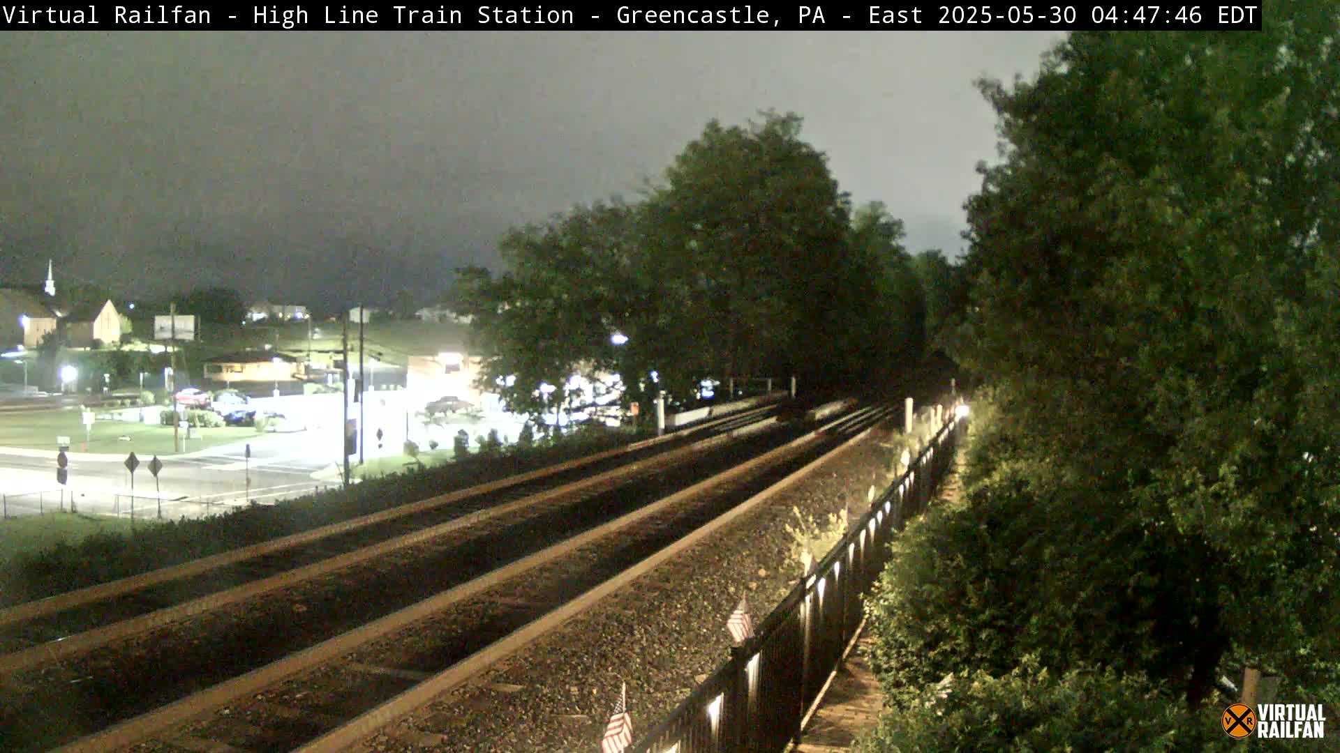 A nighttime view of train tracks extending into the distance, flanked by lush greenery and dimly lit buildings in the background under a cloudy sky.