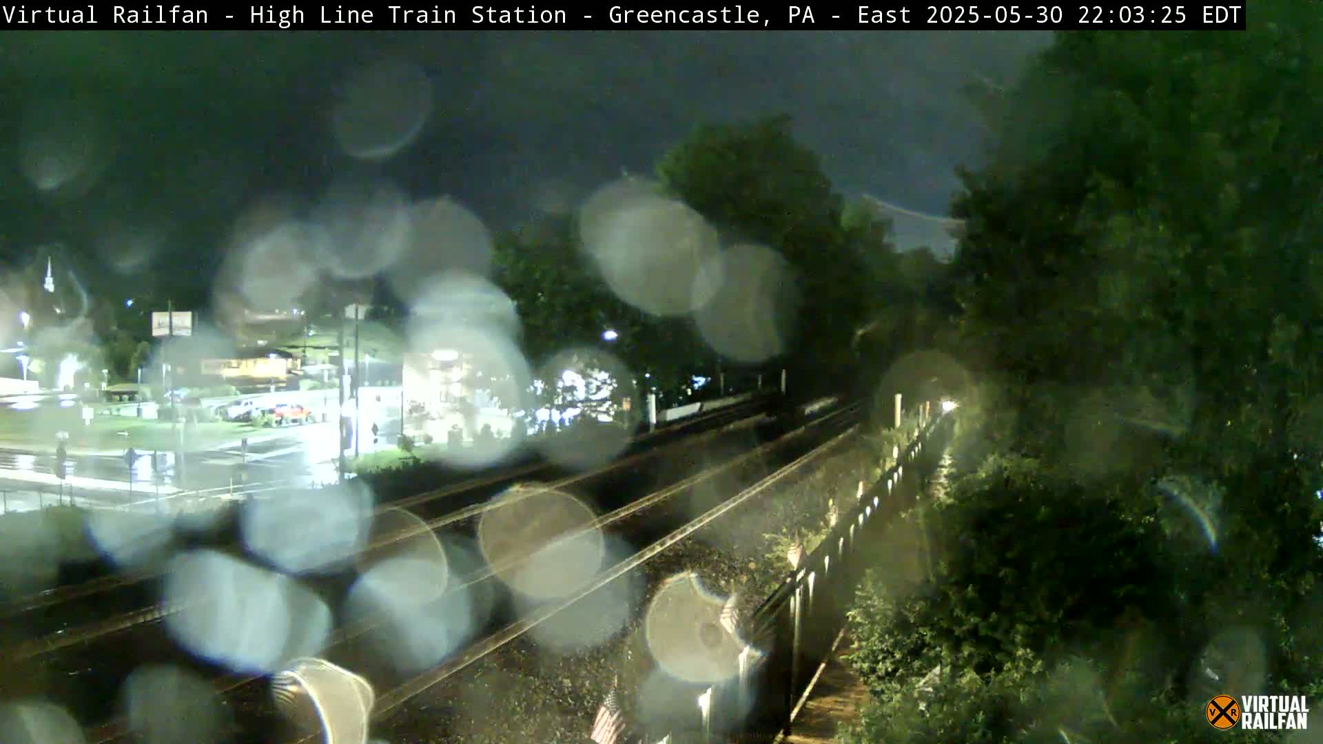 Rain-streaked nighttime view of railroad tracks near a road intersection and lush greenery.