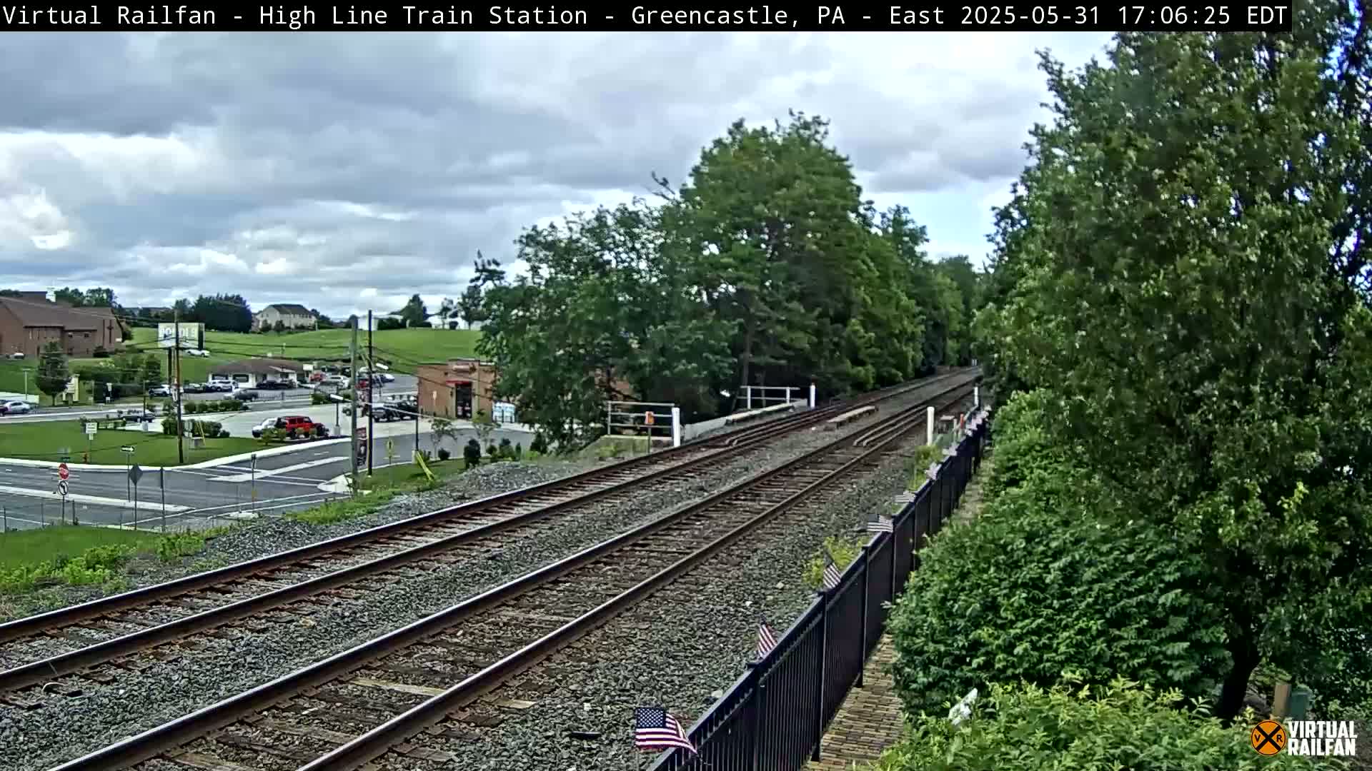 A view of train tracks extending into the distance on an overcast day, with lush green trees and a road with buildings in the background.