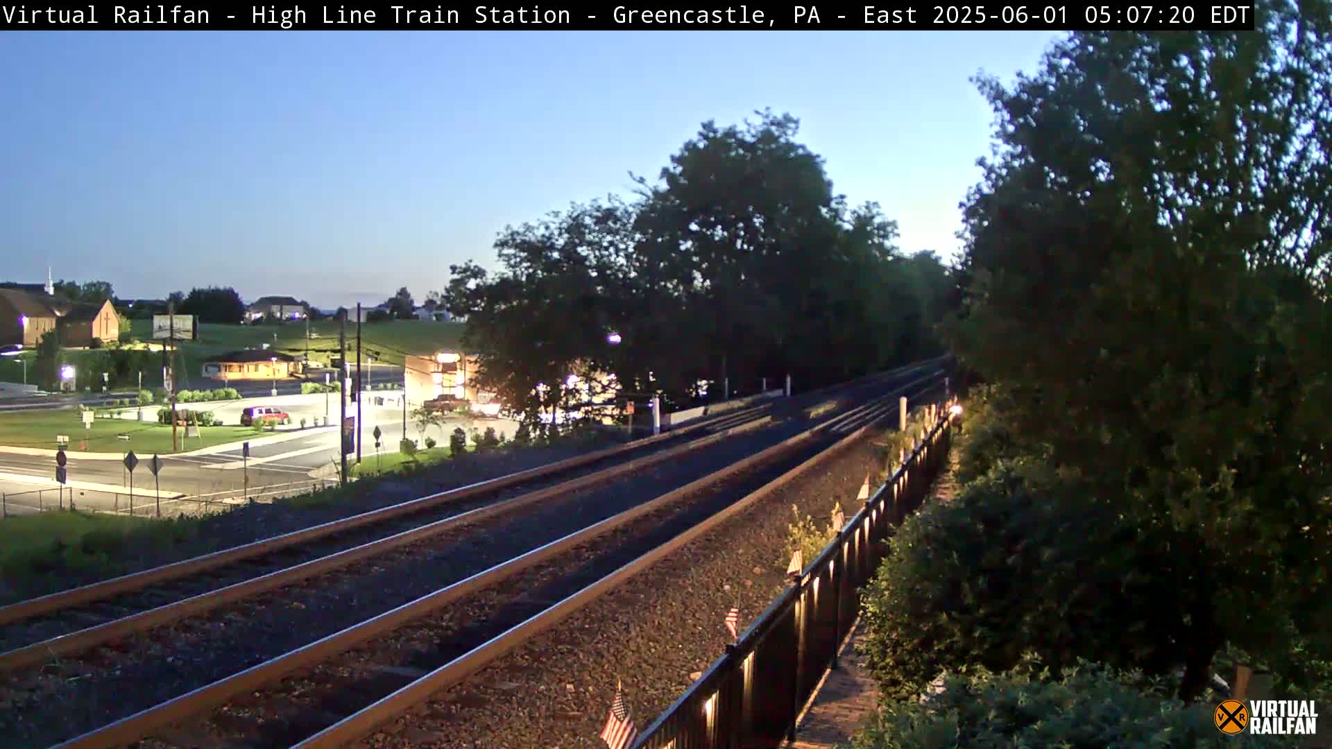 Under a twilight sky, a view of a railway track extends from the foreground into the distance, passing through a landscape of trees and a small town with illuminated buildings.
