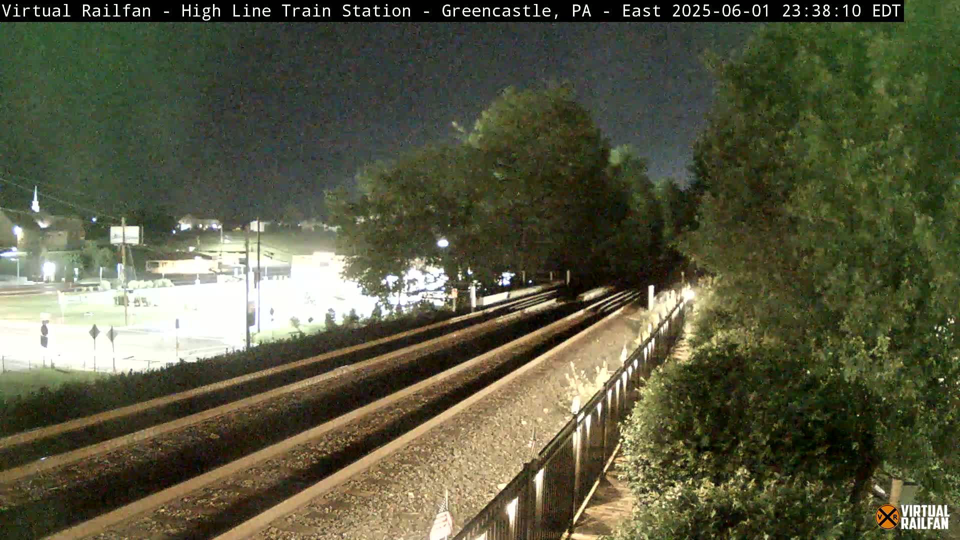 At night, three sets of train tracks extend into the distance, bordered by a fence and lush green vegetation under a dark sky.