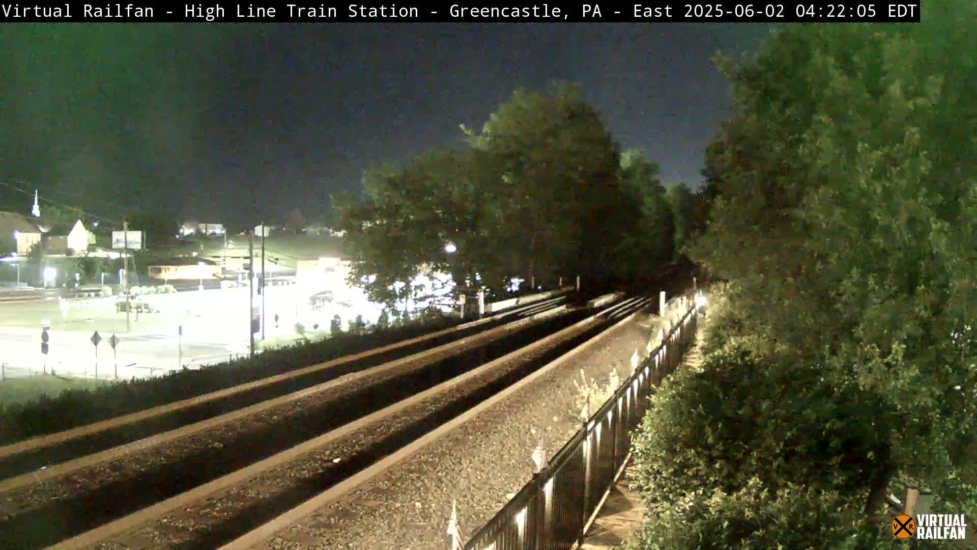A nighttime view of train tracks extending into the distance, bordered by a fence and lush greenery under a clear, dark sky.