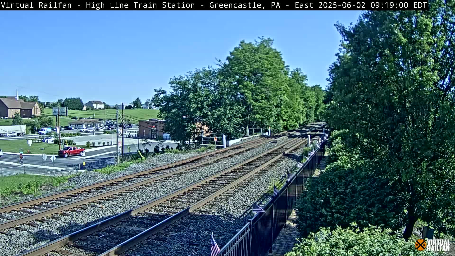 A sunny day view of railroad tracks crossing a road, with cars and buildings visible in the background and lush green trees on either side.