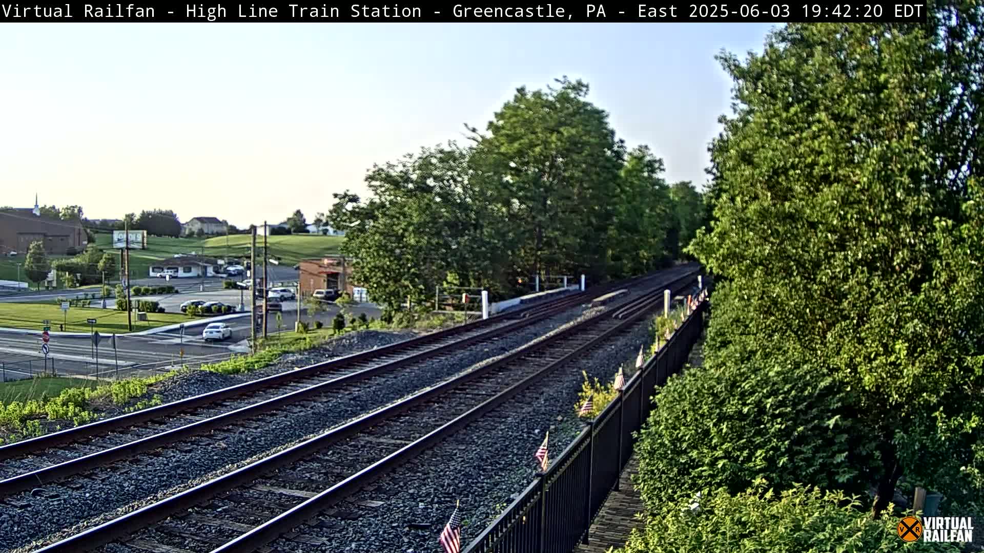 Train tracks extend into the distance under a clear evening sky, bordered by lush green vegetation and a road with cars visible in the background.