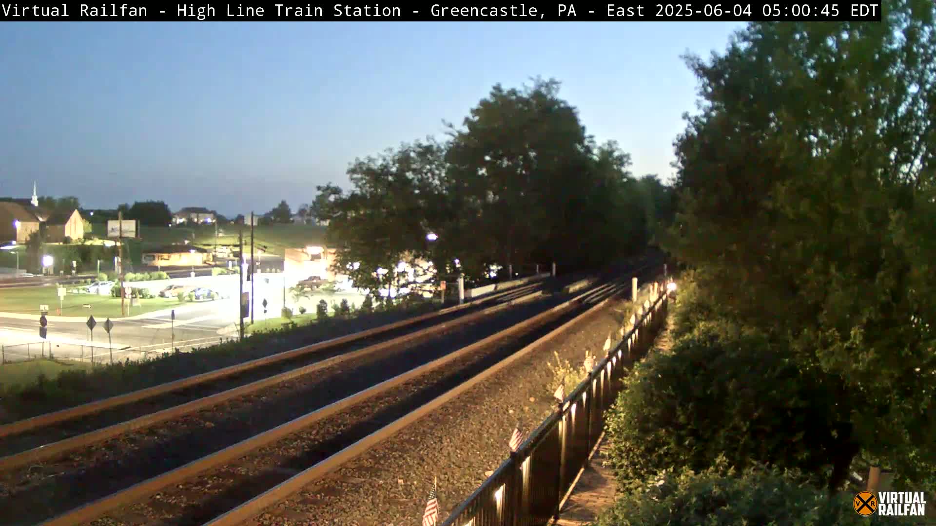 Train tracks stretch into the distance under a twilight sky, bordered by a fence and lush greenery;  a small town is visible in the background.