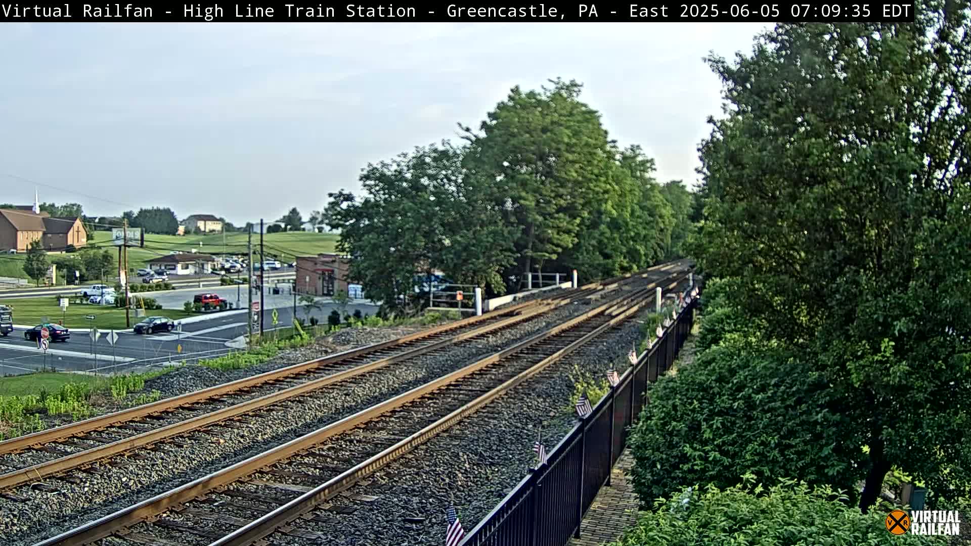A view of train tracks extending into the distance under a partly cloudy sky, with a road and buildings visible in the background.