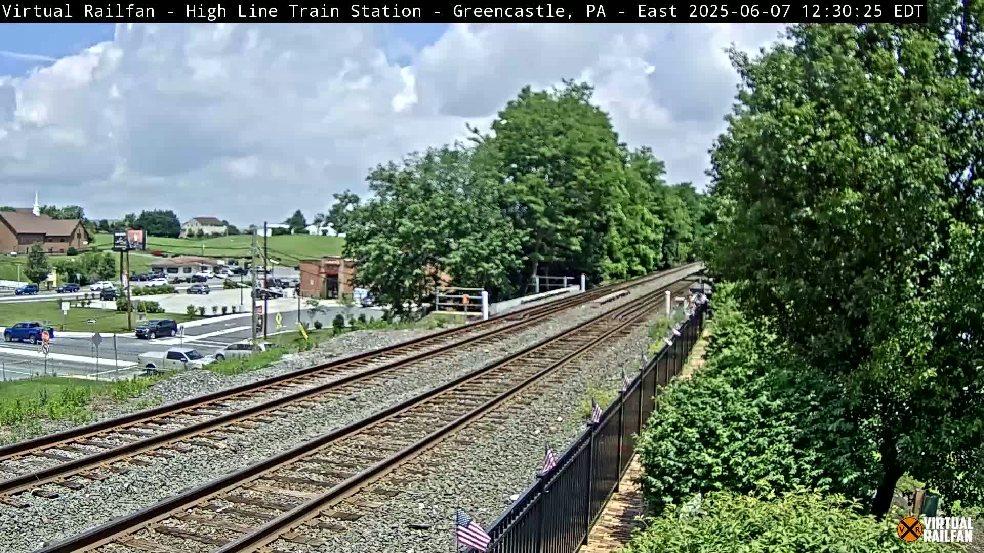 A mostly sunny day shows a view of train tracks extending into the distance, past a black fence lined with small American flags, toward a small town with buildings and cars visible in the background.