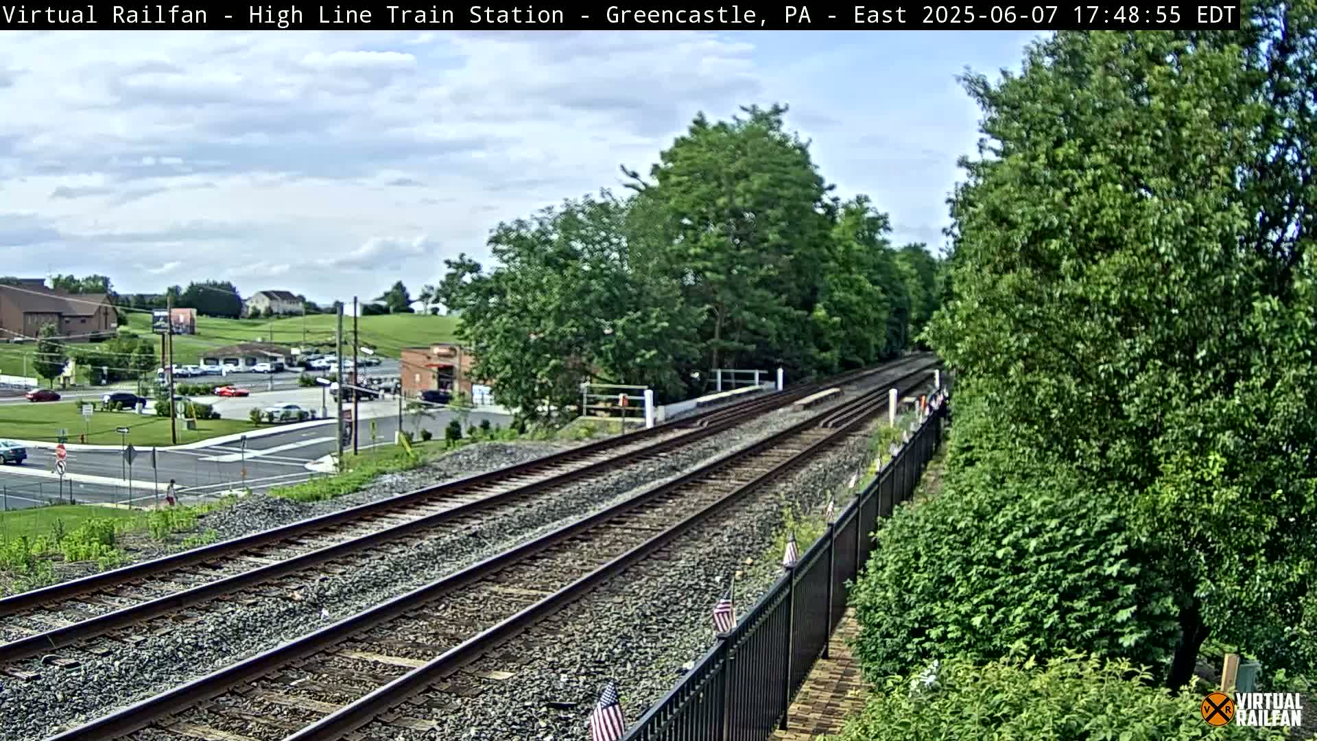 Train tracks extend into the distance under a partly cloudy sky, passing through a lush green landscape with a small town visible in the background.