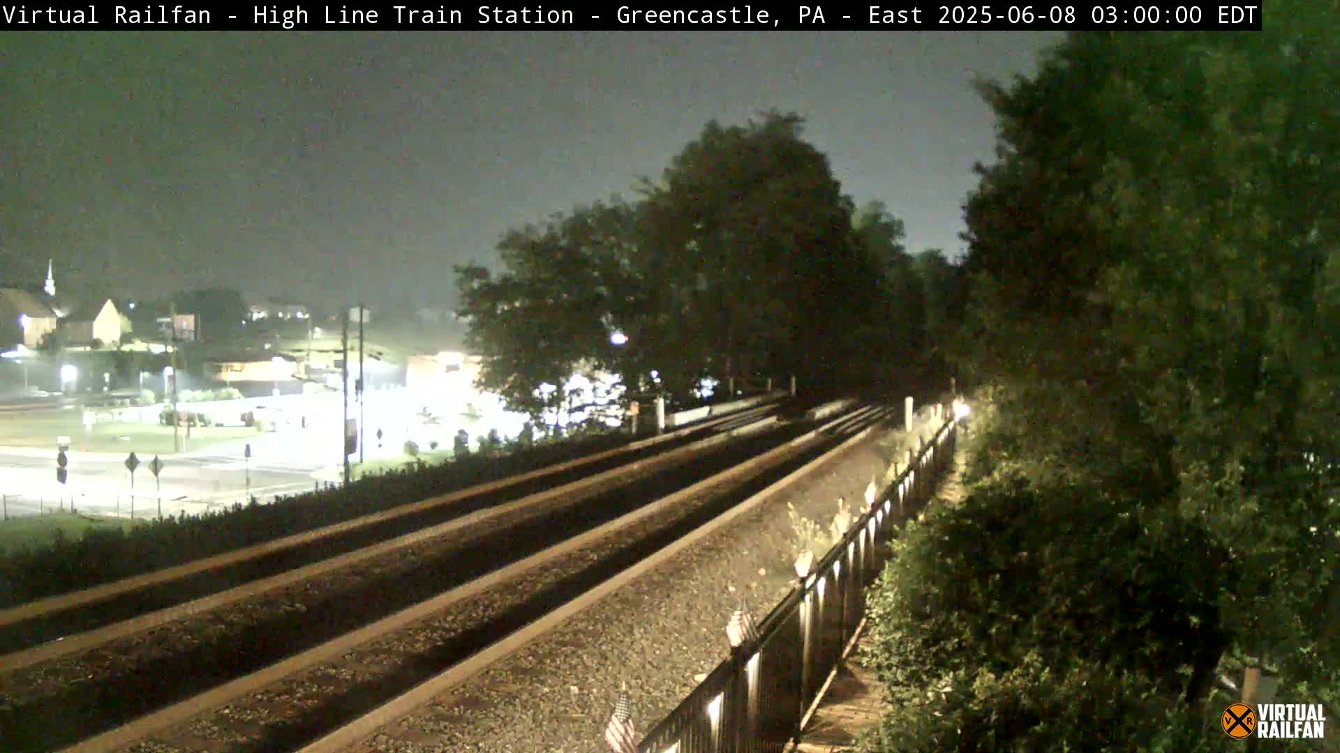 A nighttime view of train tracks extending into the distance, alongside a road and some buildings under a dark, overcast sky.