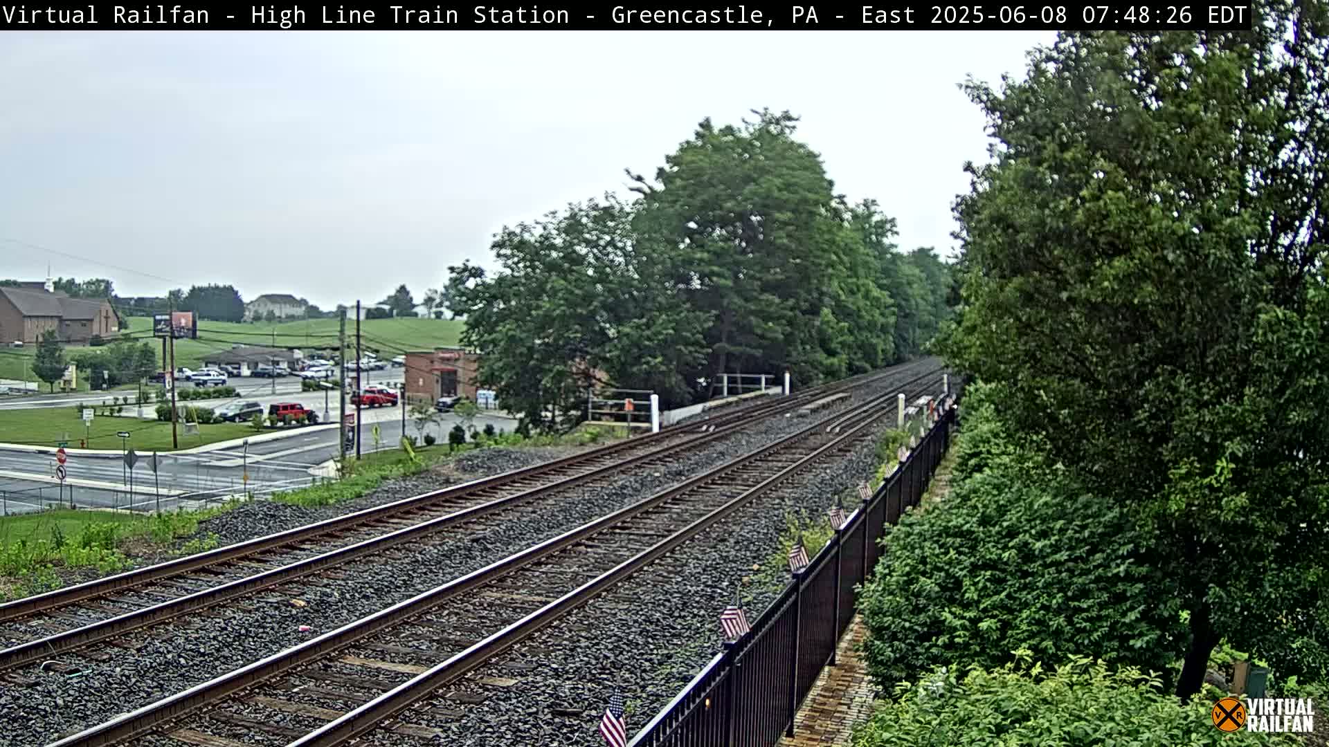 Here is a description of the image:

A view of a train track that extends into the distance, bordered by lush green trees and a black fence, on an overcast day.