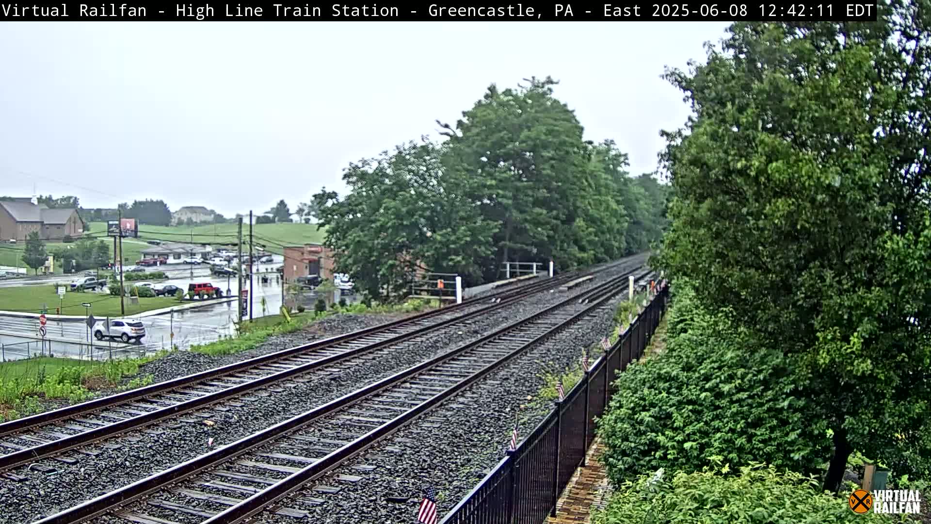 A view of a train track extending into the distance on a rainy day, with green trees and a parking lot visible in the background.