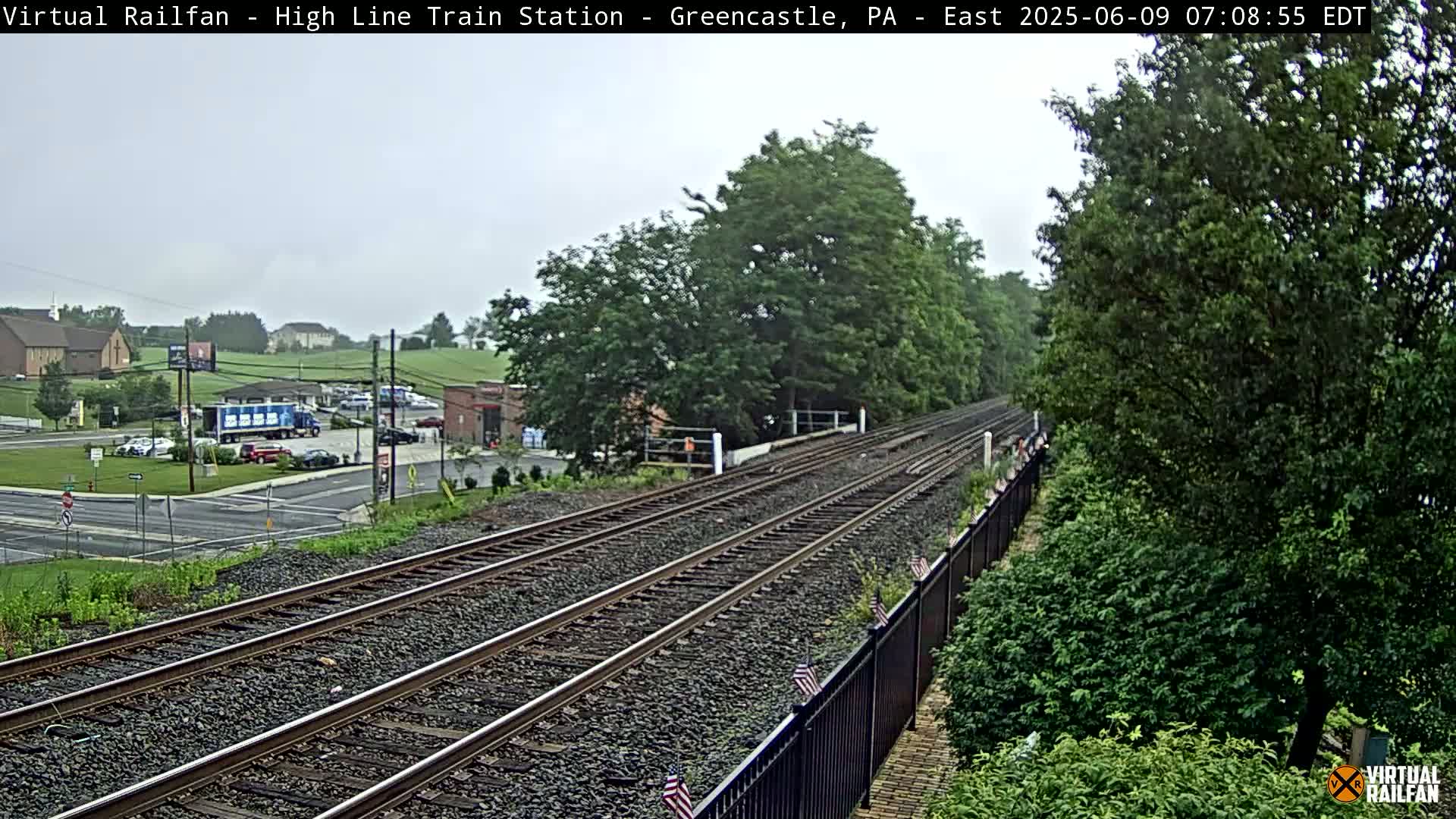 A view of railroad tracks extending into the distance on an overcast day, bordered by lush green trees and a black metal fence, with a parking lot and buildings visible in the background.