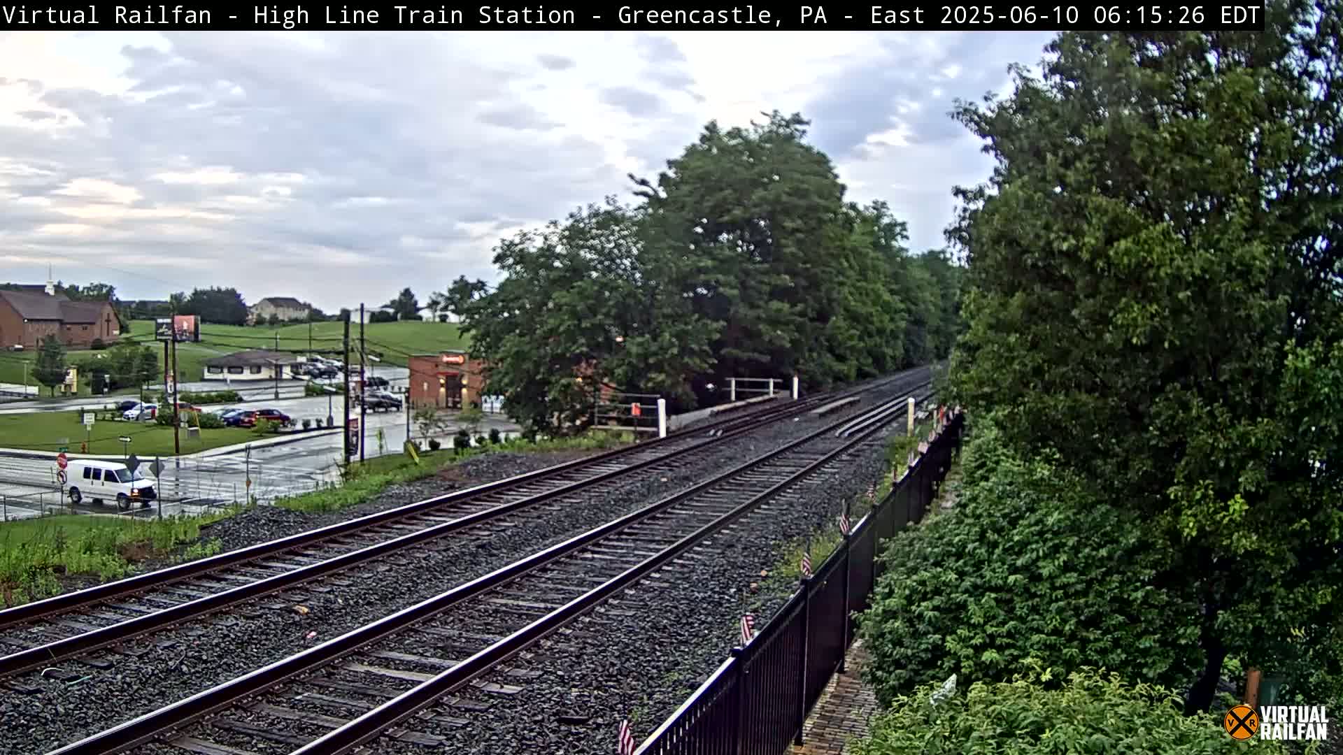A long stretch of train tracks extends into the distance, passing through a landscape of trees and a parking lot with a few cars under a partly cloudy sky.