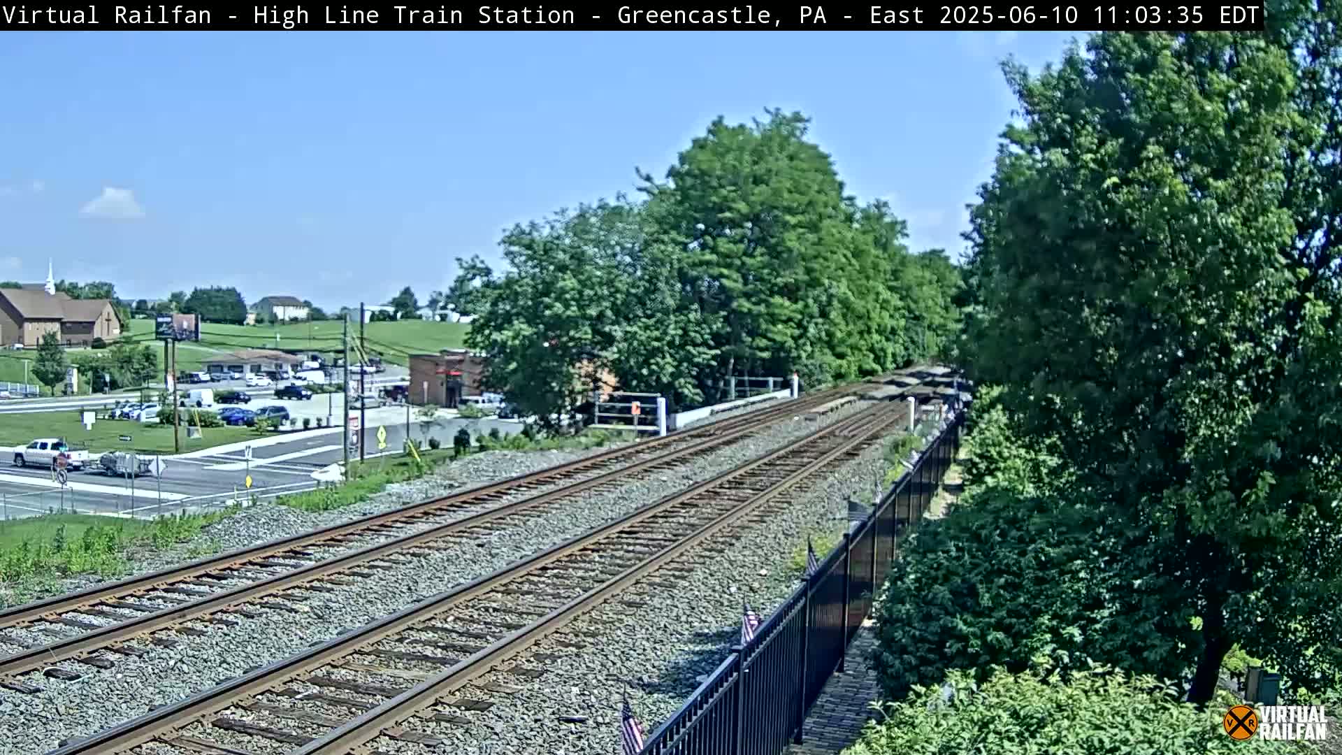 A sunny day shows a view of a train track extending into the distance, bordered by lush green trees and a black metal fence, with a parking lot and buildings visible in the background.