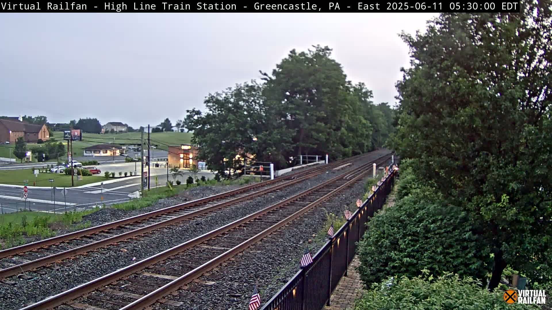 Two sets of train tracks extend into the distance under an overcast sky, passing by a small town with buildings and cars visible in the background.