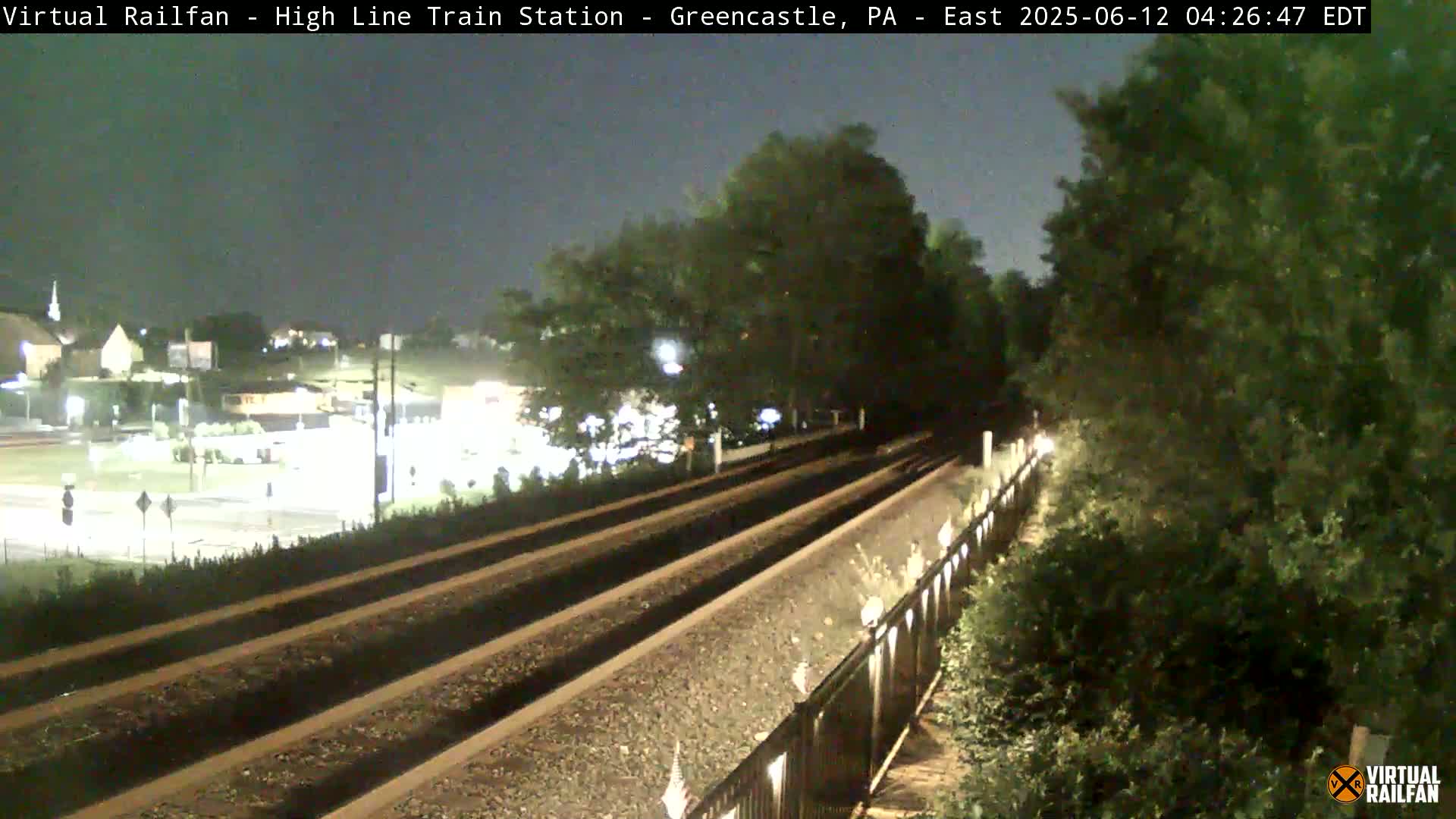 A nighttime view of train tracks extending into the distance, bordered by lush greenery, with a road and buildings visible in the background under a dark sky.