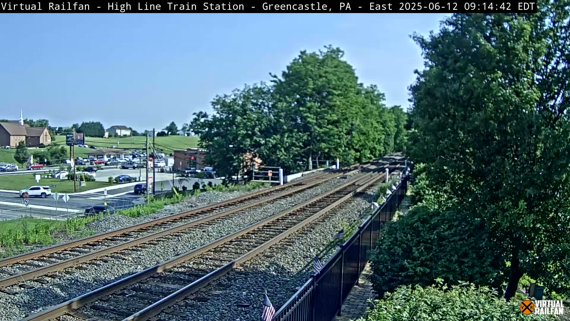 A sunny day view of train tracks stretching into the distance, alongside a road with cars parked and driving by, and lush green trees and buildings in the background.