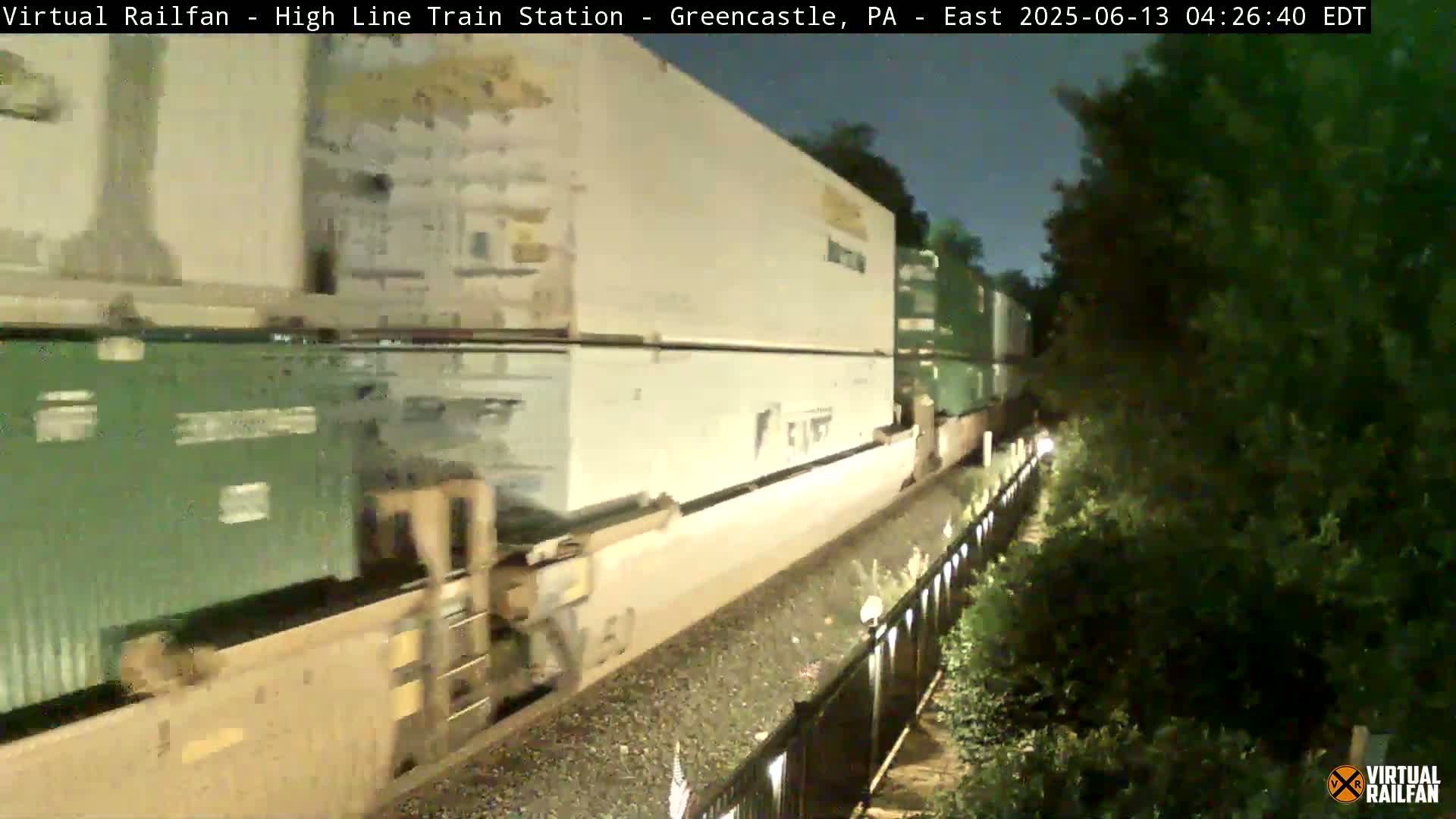 A long freight train carrying multiple shipping containers moves along a track at night.