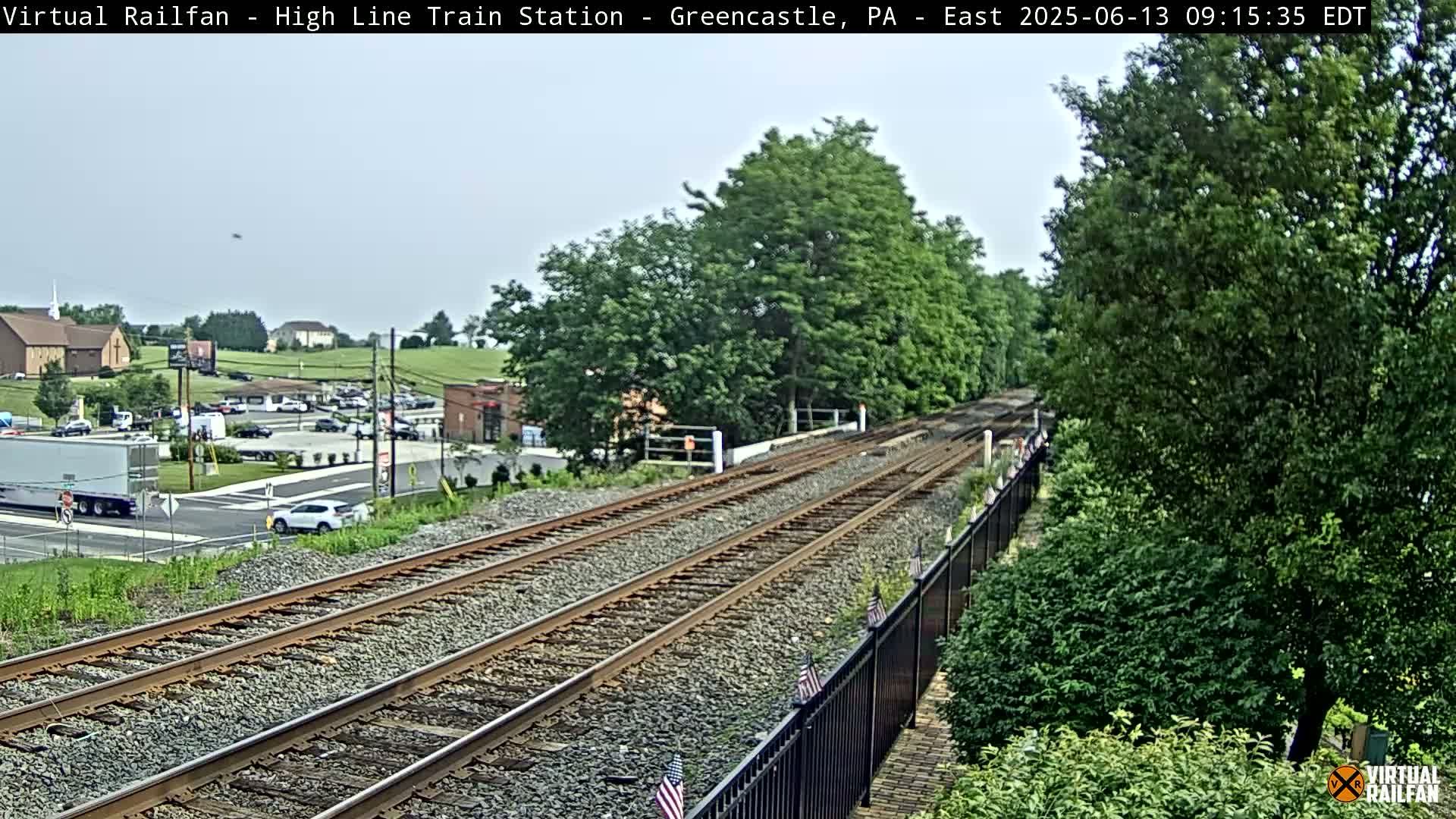 A mostly clear sky overlooks railroad tracks that extend into the distance, passing through a lush green landscape and beside a roadway with cars and buildings.
